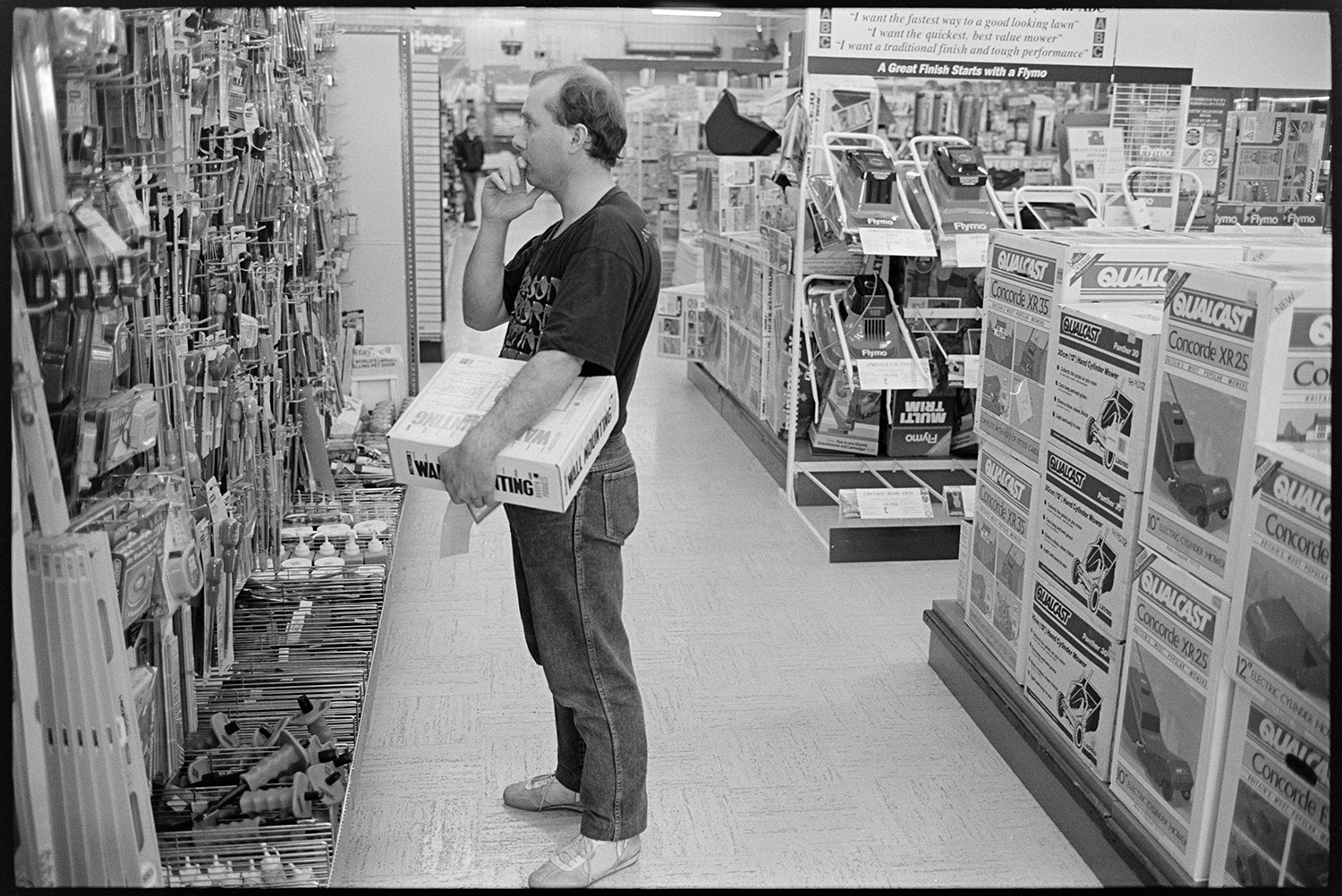 Customer selecting goods from shelves of DIY supermarket. 
[A man looking at goods for sale in a DIY shop in Barnstaple. Lawn mowers are on display behind him.]