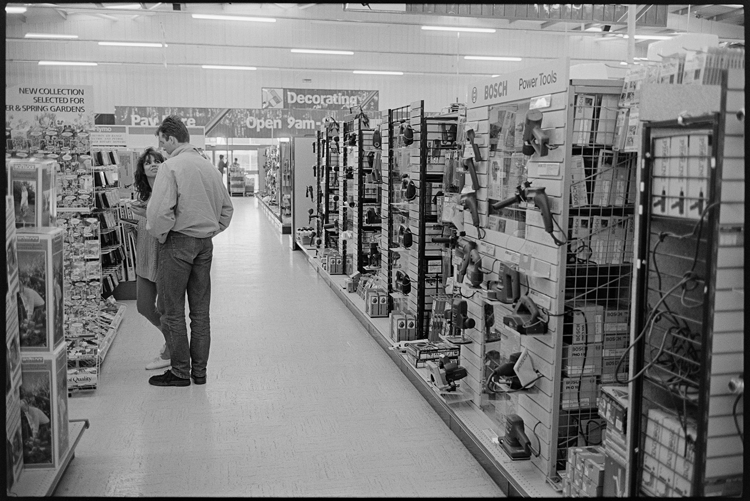 Customer selecting goods from shelves of DIY supermarket. 
[A man and woman looking at goods for sale in a DIY shop in Barnstaple. Goods, including power tools and seeds, are displayed for sale on the shelves.]