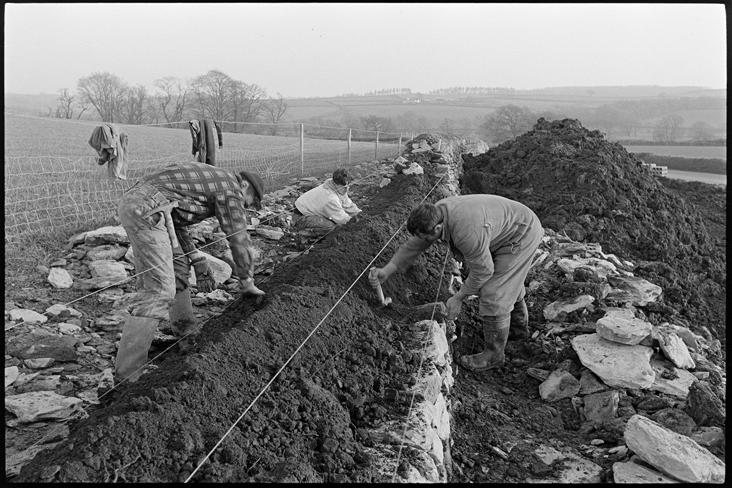 Men building dry stone wall beside road. 
[Three men building a dry stone wall at Leigh Cross, Chulmleigh. Their jackets are hung over a wire fence by the adjacent field and a large mound of earth can be seen in the background.]