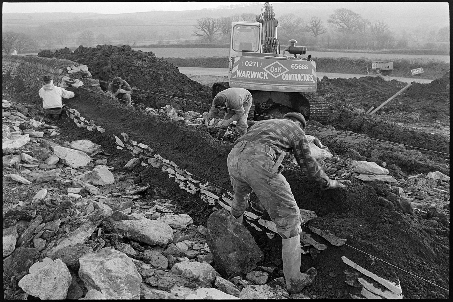 Men building dry stone wall beside road. 
[Four men building a dry stone wall at Leigh Cross, Chulmleigh. A digger and large mound of earth can be seen in the background.]