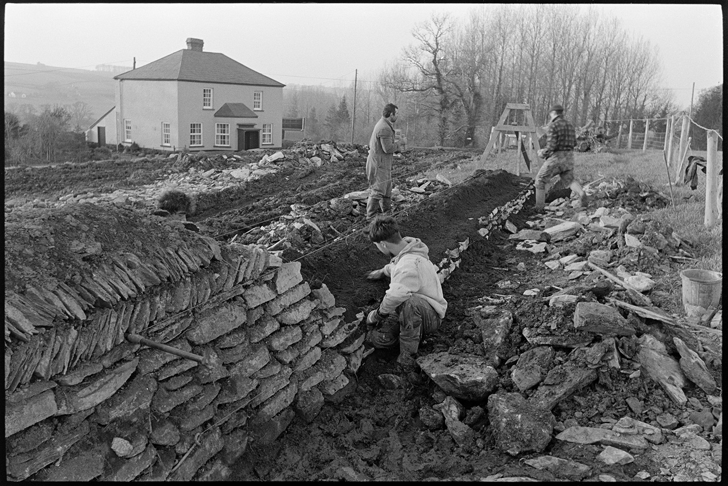 Men building dry stone wall beside road. 
[Four men building a dry stone wall at Leigh Cross, Chulmleigh. A house can be seen in the background.]