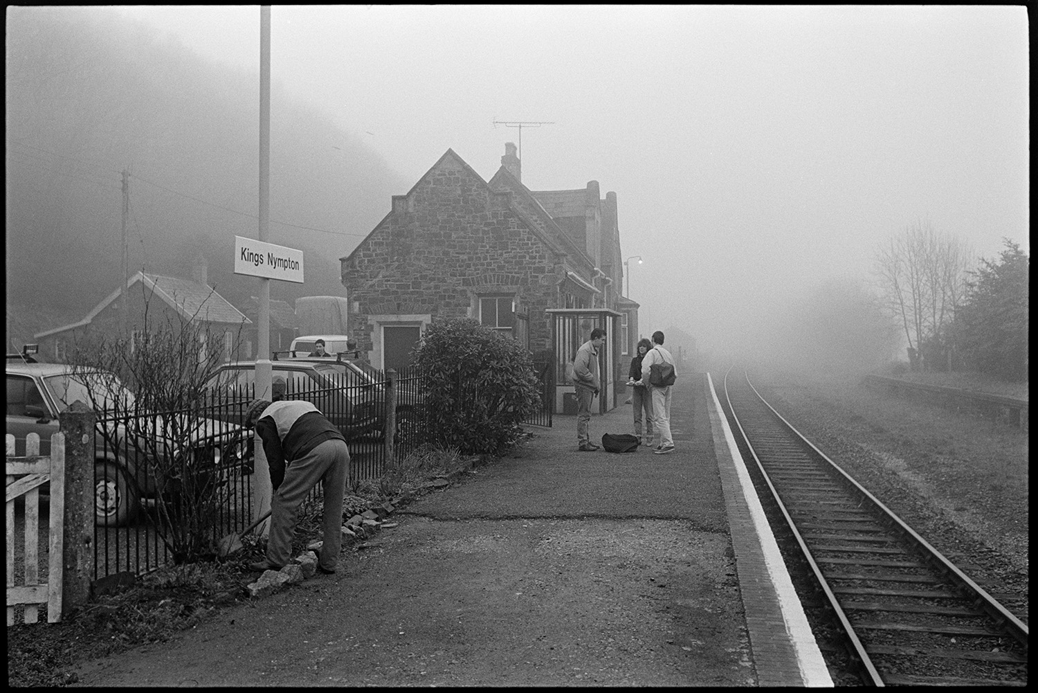 Railway station, platform with train and passengers. 
[Passengers waiting on the platform at Kings Nympton railway station. Parked cars can be seen in the car park and the track and station building are visible.]