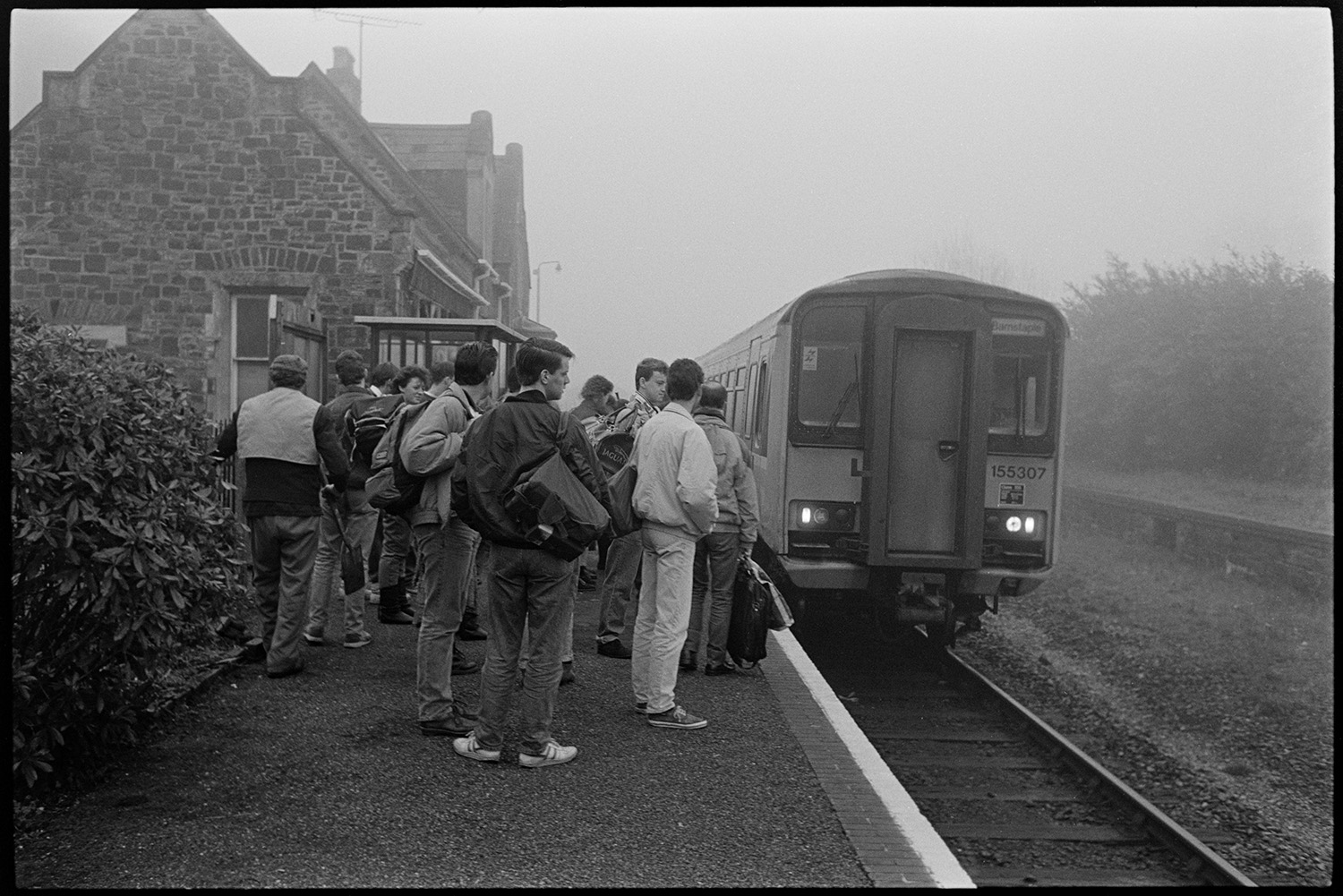 Railway station, platform with train and passengers. 
[Passengers waiting on the platform at Kings Nympton railway station to board at train which has just pulled in. The station buildings is visible in the background.]