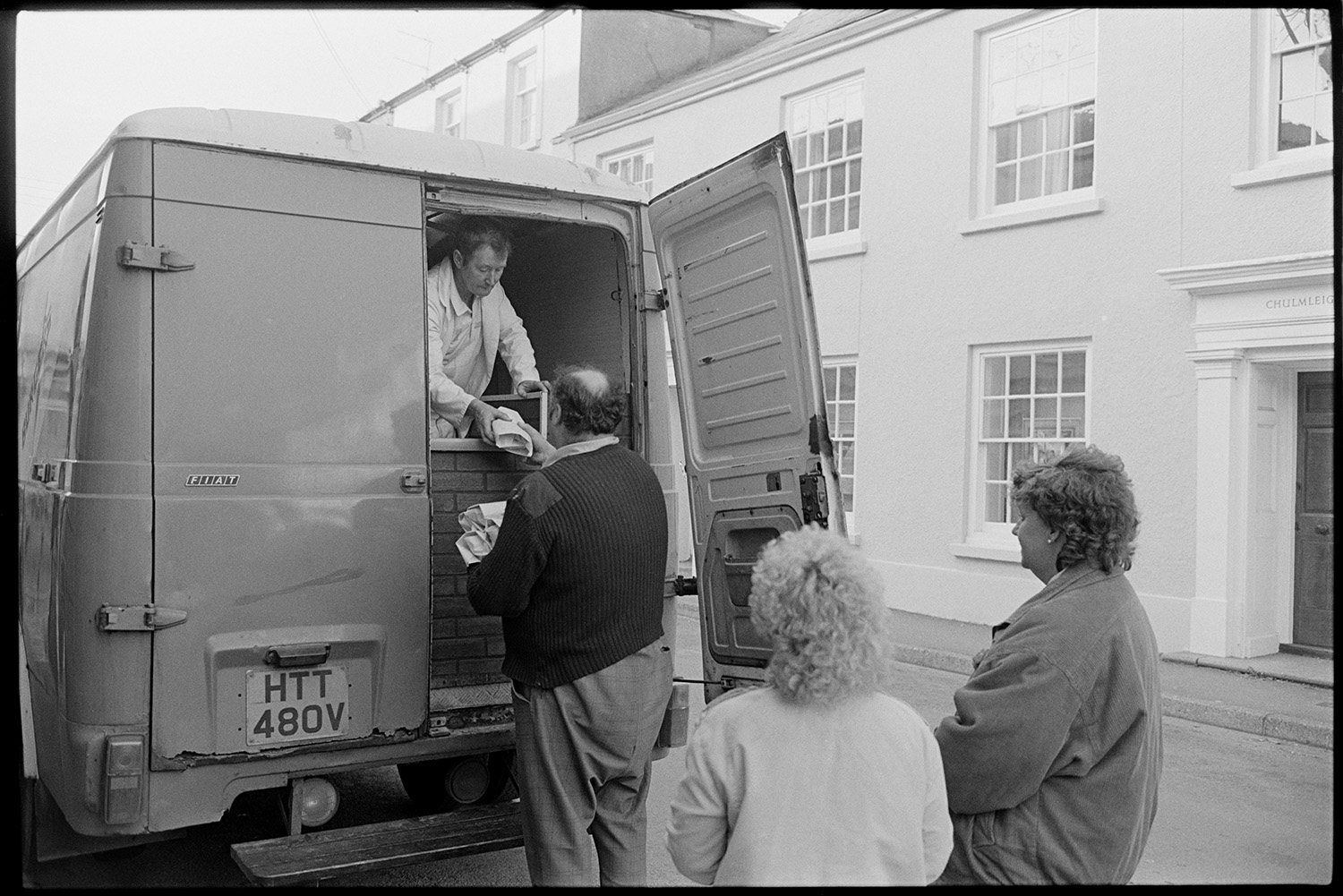 Fish and Chip van serving people in village street. 
[Men and women queuing up to buy fish and chips from a van, in a street in Chulmleigh.]