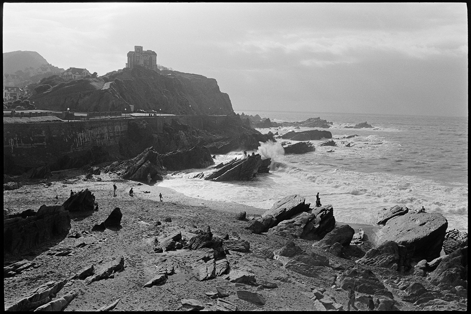 People walking on rocky beach with stormy sea and sky, clouds. 
[People walking between rocks on the beach at Ilfracombe. Waves are crashing over the rocks and a building is visible on the clifftop in the background, against a  cloudy sky.]