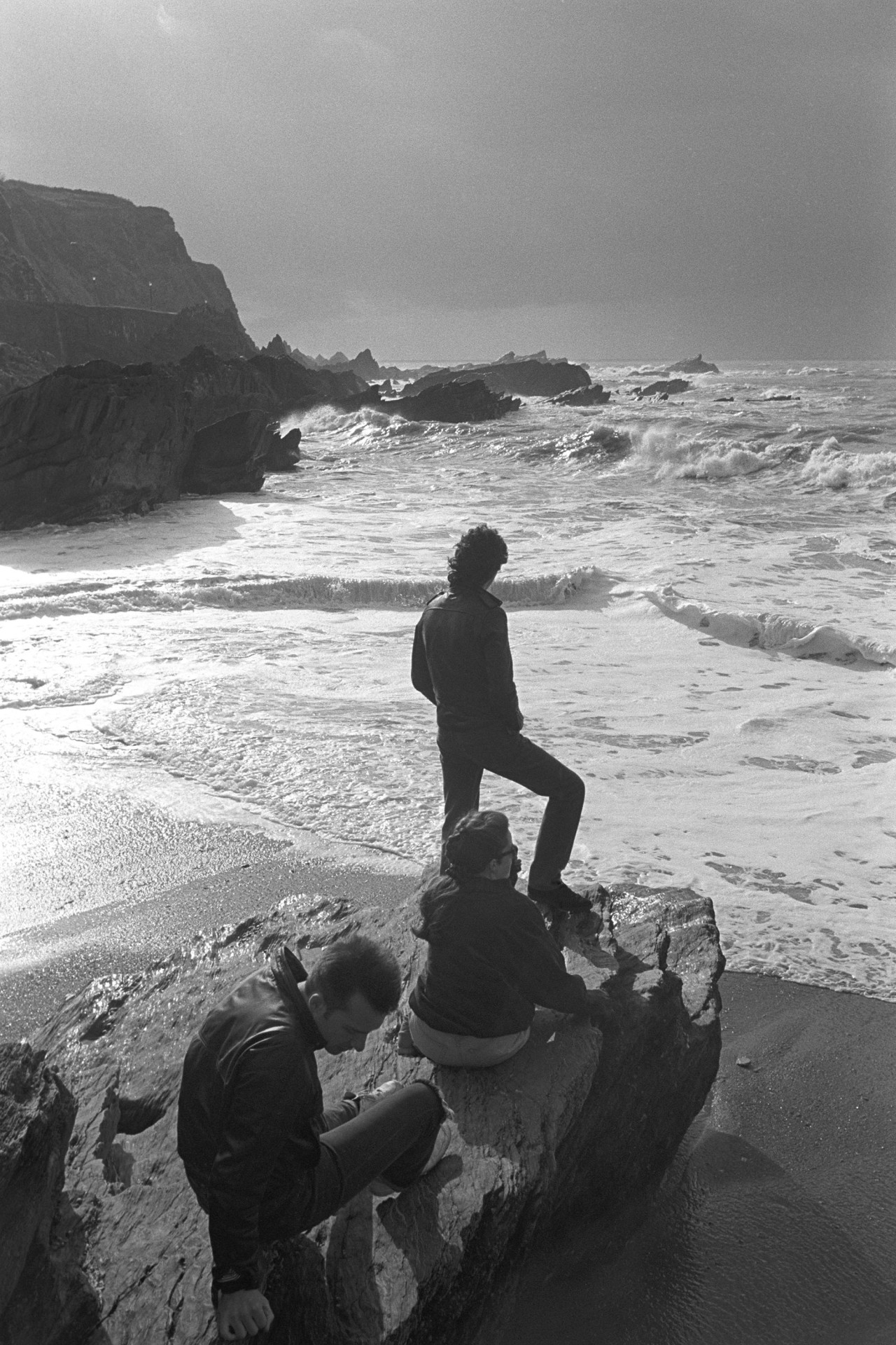 Youths on rock contemplating stormy sea and sky. 
[Two young men and a woman stood and sat on a large rock at Ilfracombe beach, looking out at the rough sea and stormy sky.]