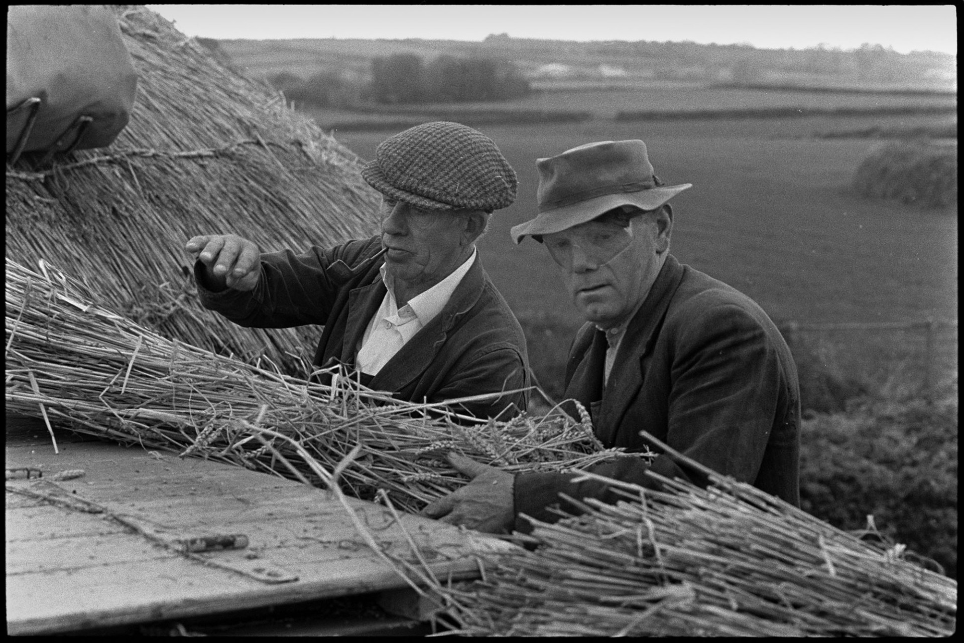 Reed combing, men working, ricks, tractors, etc. <br />
[Two men holding bundles of reed which they are feeding into a reed comber. One man is smoking a pipe and the other man is wearing an eye mask. A thatched wheat rick can be seen in the background.]