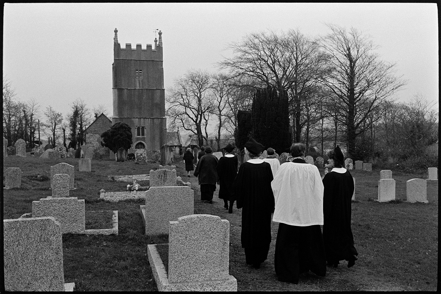 Christening with Bishop at village church, choir, uniform.
[Group of clergy and members of the congregation walking along a path between gravestones towards Inwardleigh Church Tower. Leafless trees are visible behind the church.]