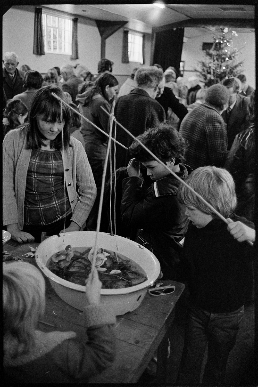 Christmas sale in village hall games and jumble.
[A woman watching three children playing a fishing game at a Christmas sale in Dolton Village Hall. A crowd at the sale and Christmas tree can be seen in the background.]