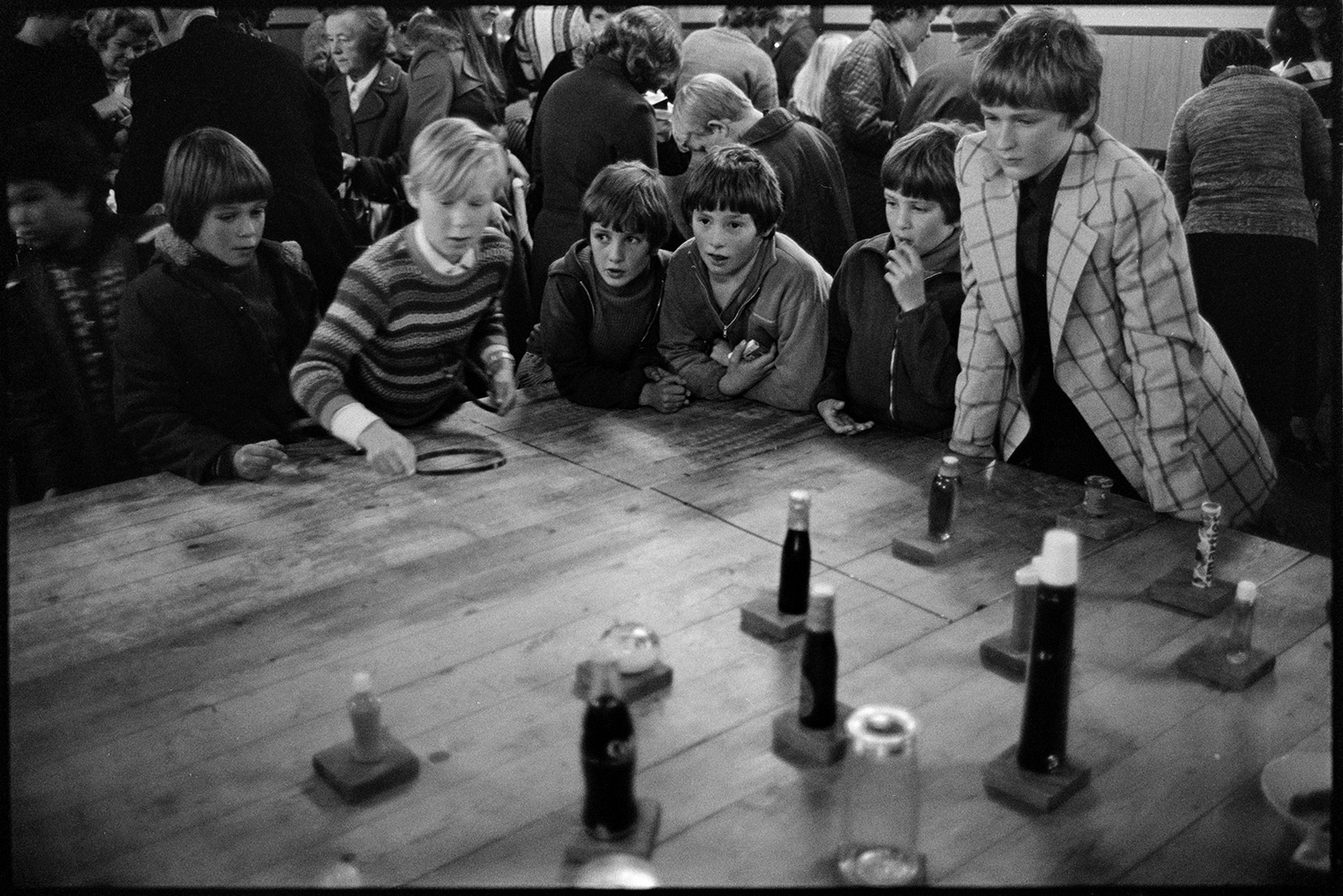 Christmas sale in village hall games and jumble.
[A group of children and watching a boy playing a hoop-la game at a Christmas sale in Dolton Village Hall. A crowd at the sale can be seen in the background.]