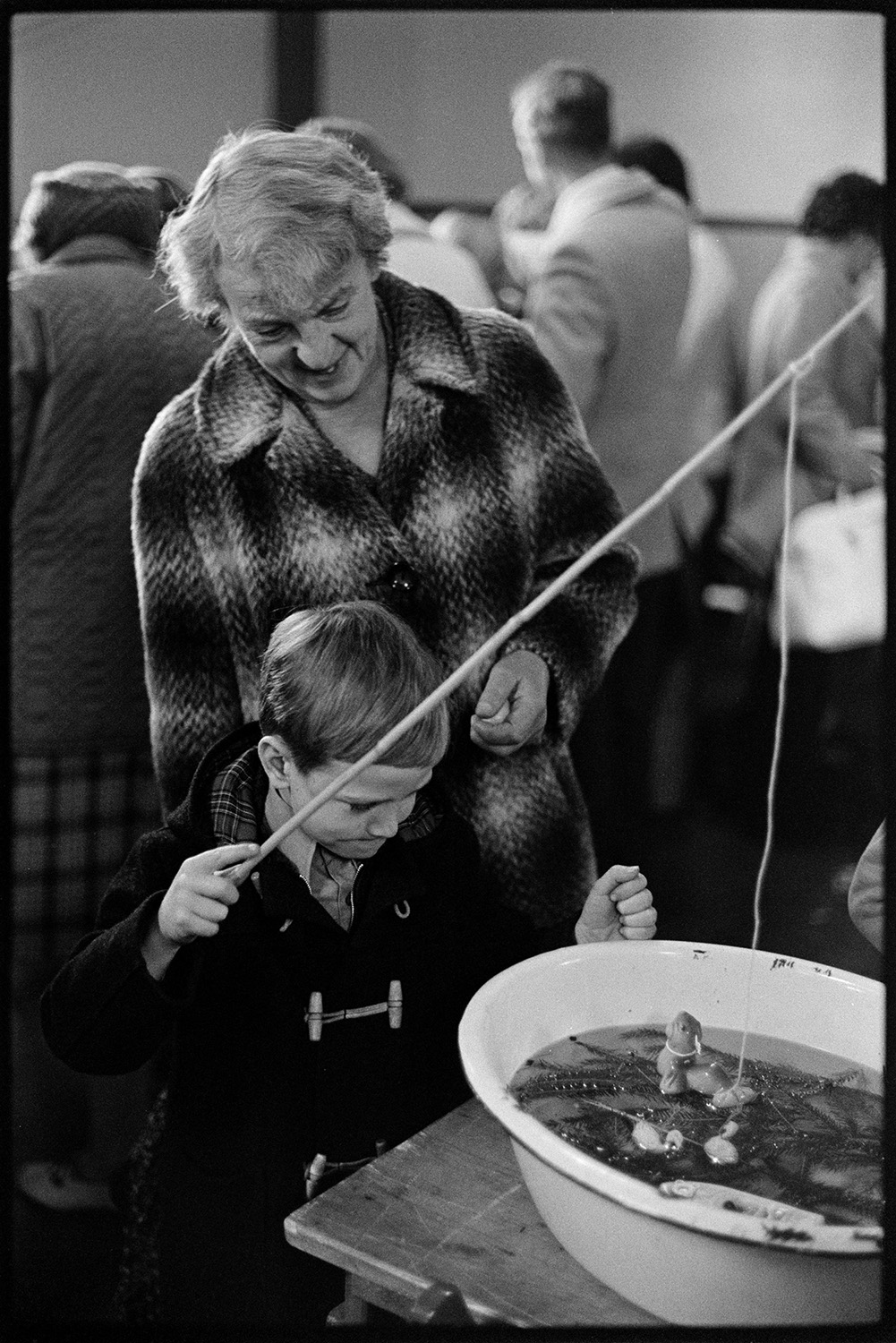 Christmas sale in village hall games and jumble.
[A woman watching a young boy playing a fishing game at a Christmas sale in Dolton Village Hall. Other people at the sale can be seen in the background.]