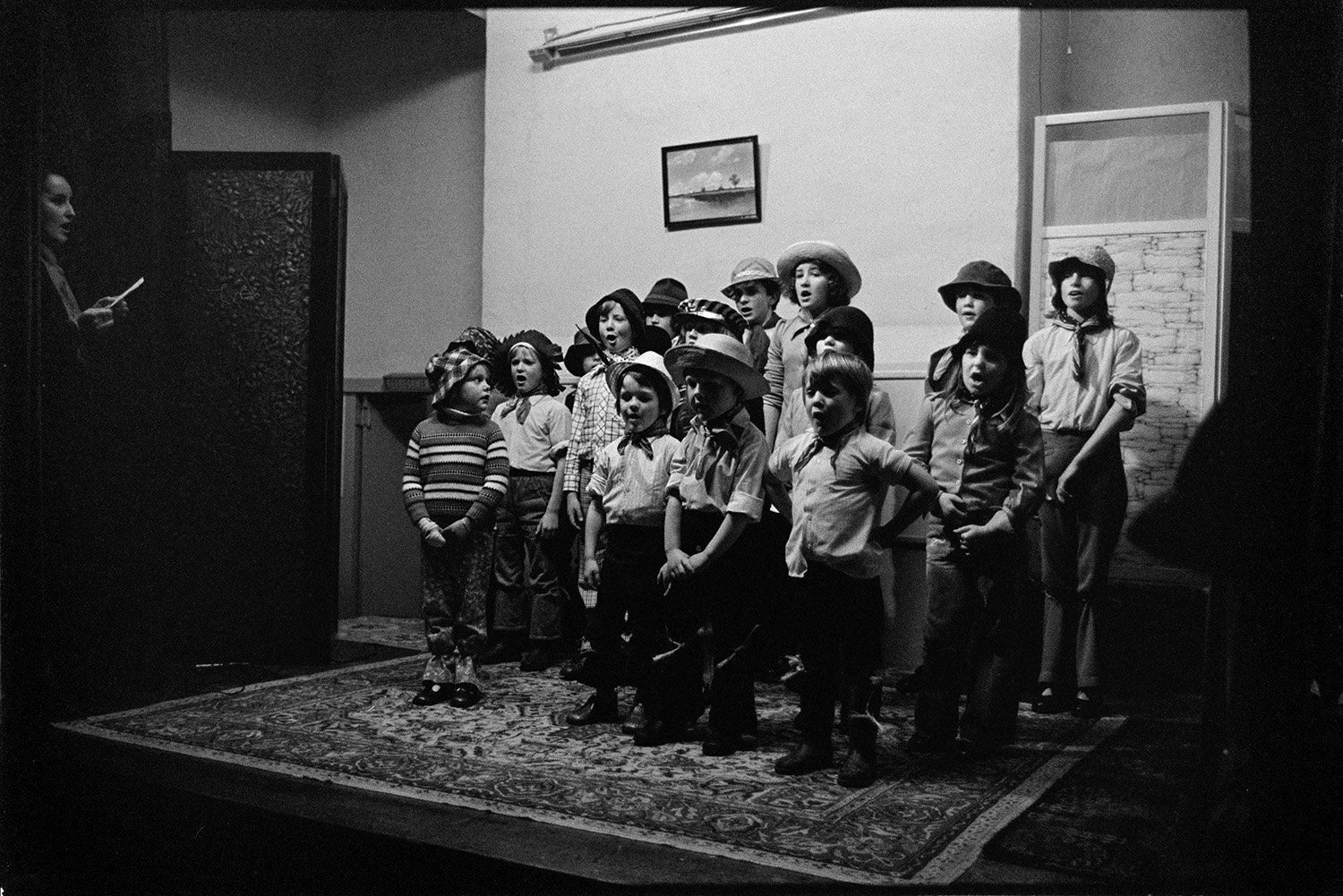 Christmas Show in village hall children's choir.
[A choir of 15 children wearing neck scarves and sun hats being led by a woman, singing in the Christmas Show in Merton Village Hall.]