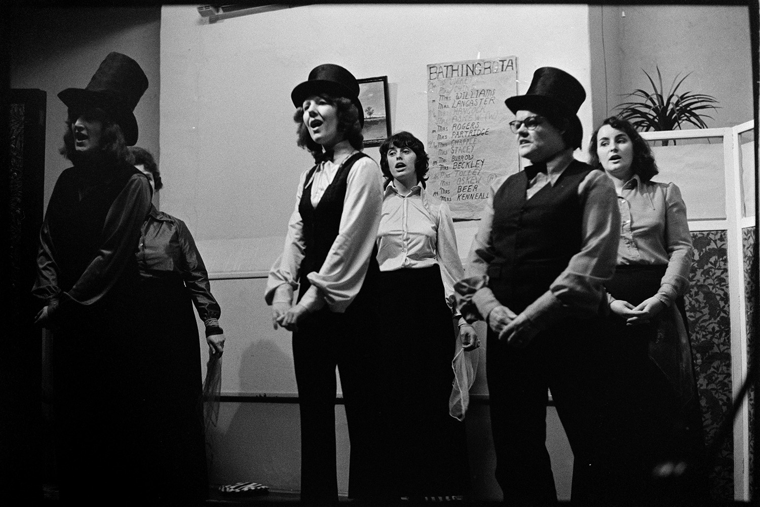 Christmas Show in village hall Musical turns.
[A group of singers, some wearing top hats, performing in the Christmas Show in Merton Village Hall. On the wall behind the singers a poster with a 'Bathing Rota' can be seen.]
