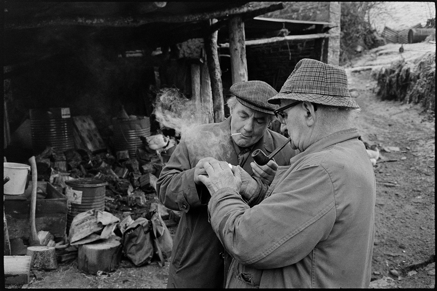 Wooden sheds with two farmers.
[Archie Parkhouse and Ivor Brock at Millhams, Dolton sharing a flame to light a cigarette and pipe. They are standing in front of a wooden open fronted shed containing equipment, storage drums, an axe and logs.]