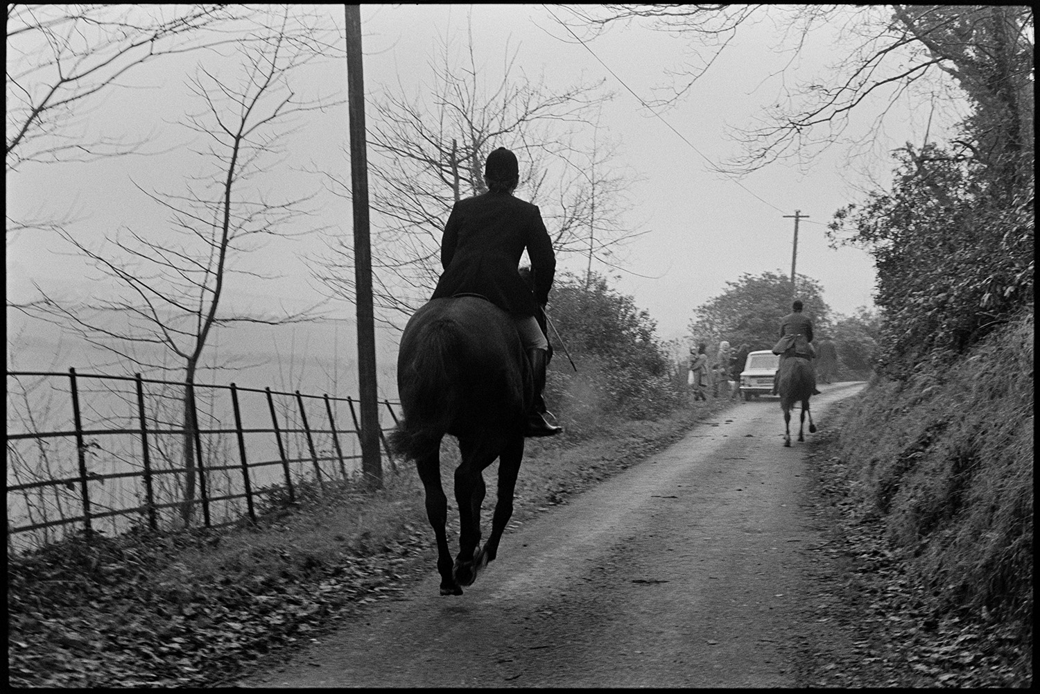 Hunt in road, waiting to move, listening for hounds. 
[Two mounted horse riders in hunting clothes riding down a lane at Halsdon, Dolton on a fox hunt. A parked car and hunt followers can be seen further down the lane.]
