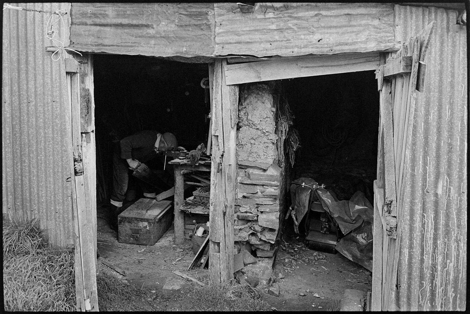 Shed door.
[A corrugated iron shed with two wooden door entrances, possibly at Dolton. Some of the stored items, including a roller or lawn mower, and a man using a saw at a work bench are visible. An interior cob wall can also be seen between the two entrances. ]