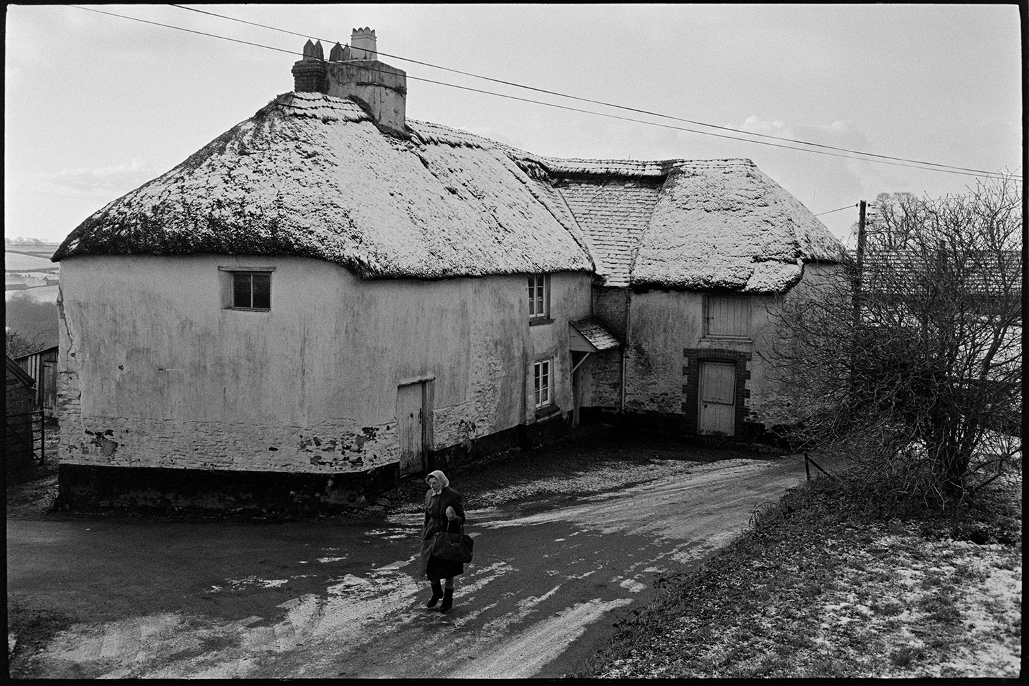 Cob and thatch farm in village in snow.
[A woman walking along an icy road in front of Elliots cob and thatched farmhouse in Roborough. The thatched roof is covered with snow.]