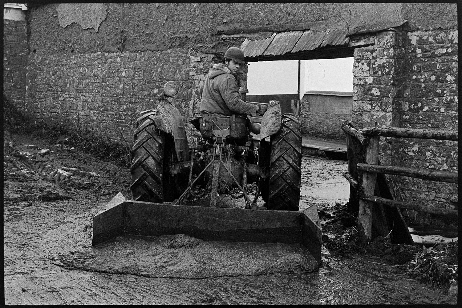 Clearing slurry with tractor and scraper.
[Graham Ward or David Ward on a tractor fitted with a scraper clearing slurry in the farmyard of Parsonage, Iddesleigh, by a stone and cob wall.]