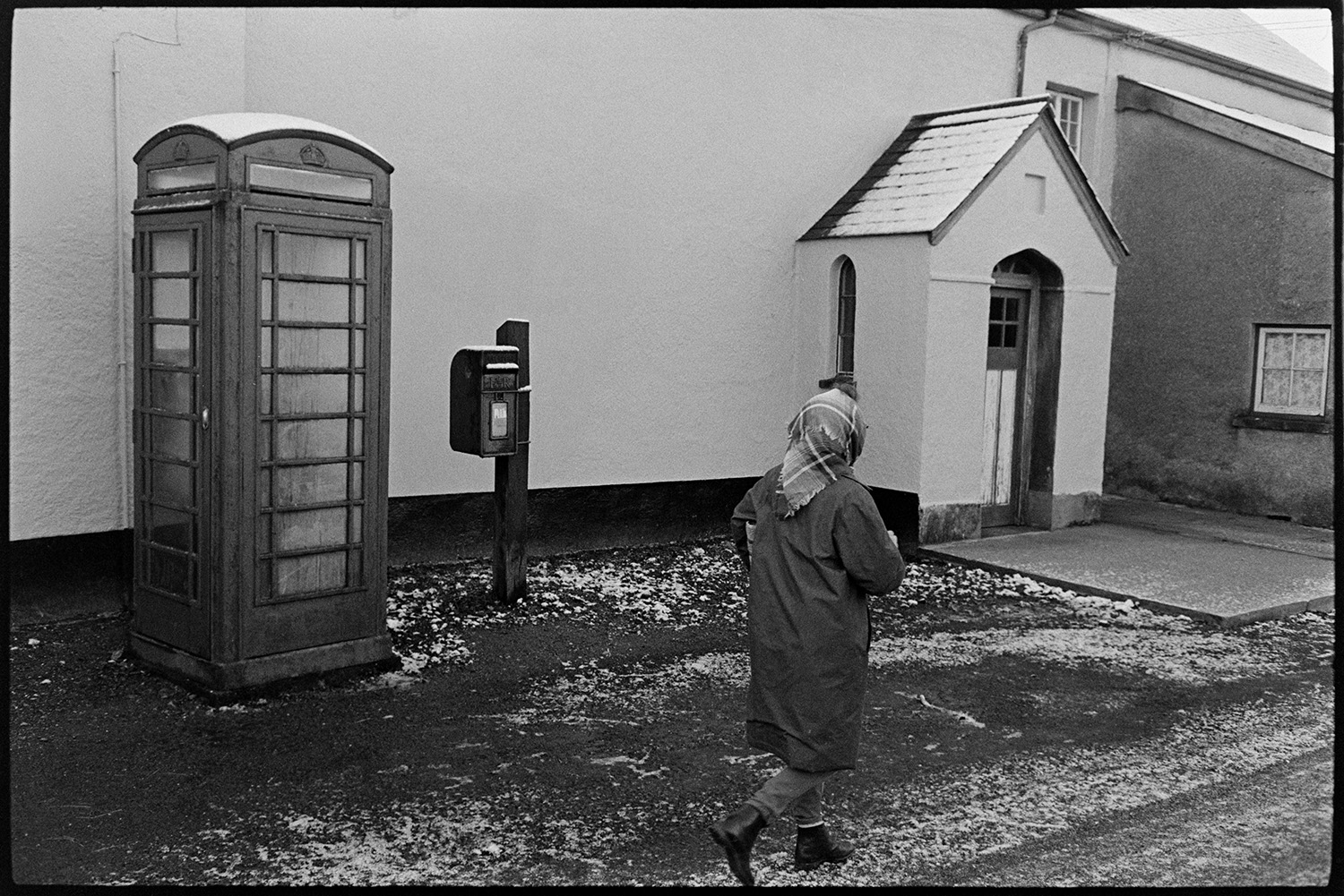 Village street with snow, telephone kiosk and letter box.
[A woman walking along an icy road past a telephone box and post box in Riddlecombe.]