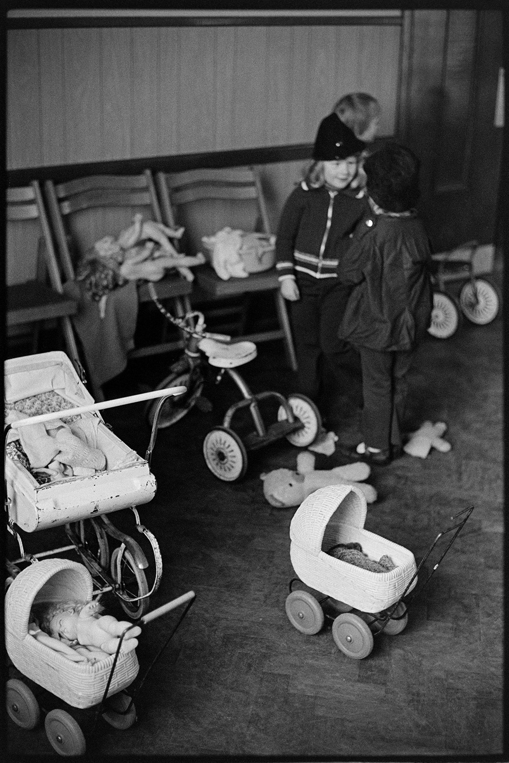 Play group toys, prams, tricycle.
[Children at a play group in Dolton Village Hall, playing with dolls, toy prams and a tricycle.]