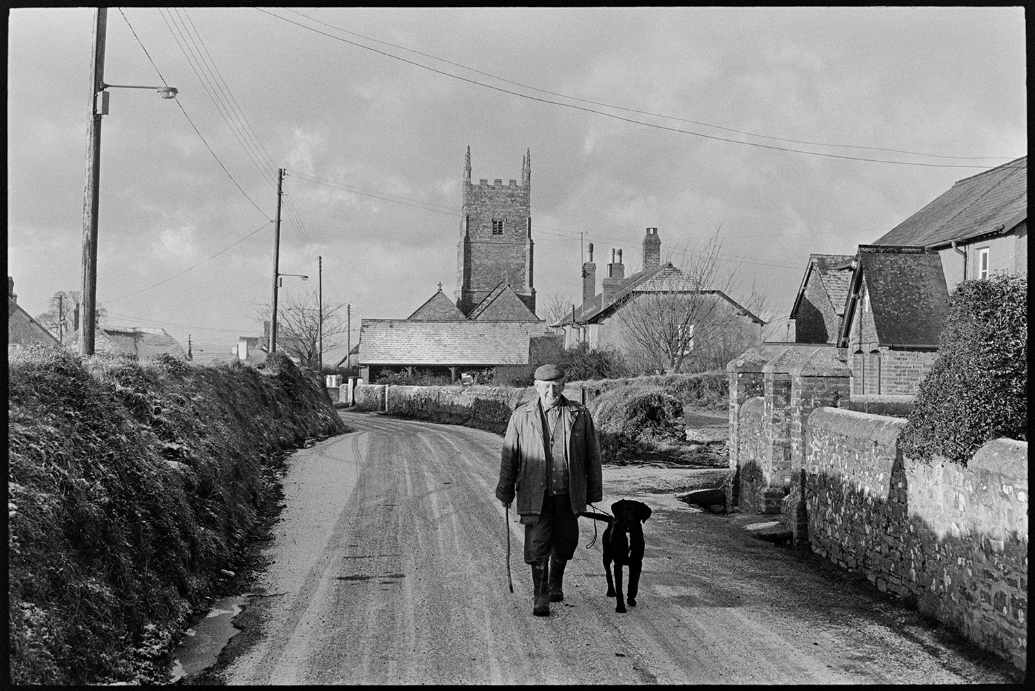Village street under snow.
[Bob Friend walking a dog along a street in Roborough which has a light covering of snow. Houses and the church tower are visible in the background.]
