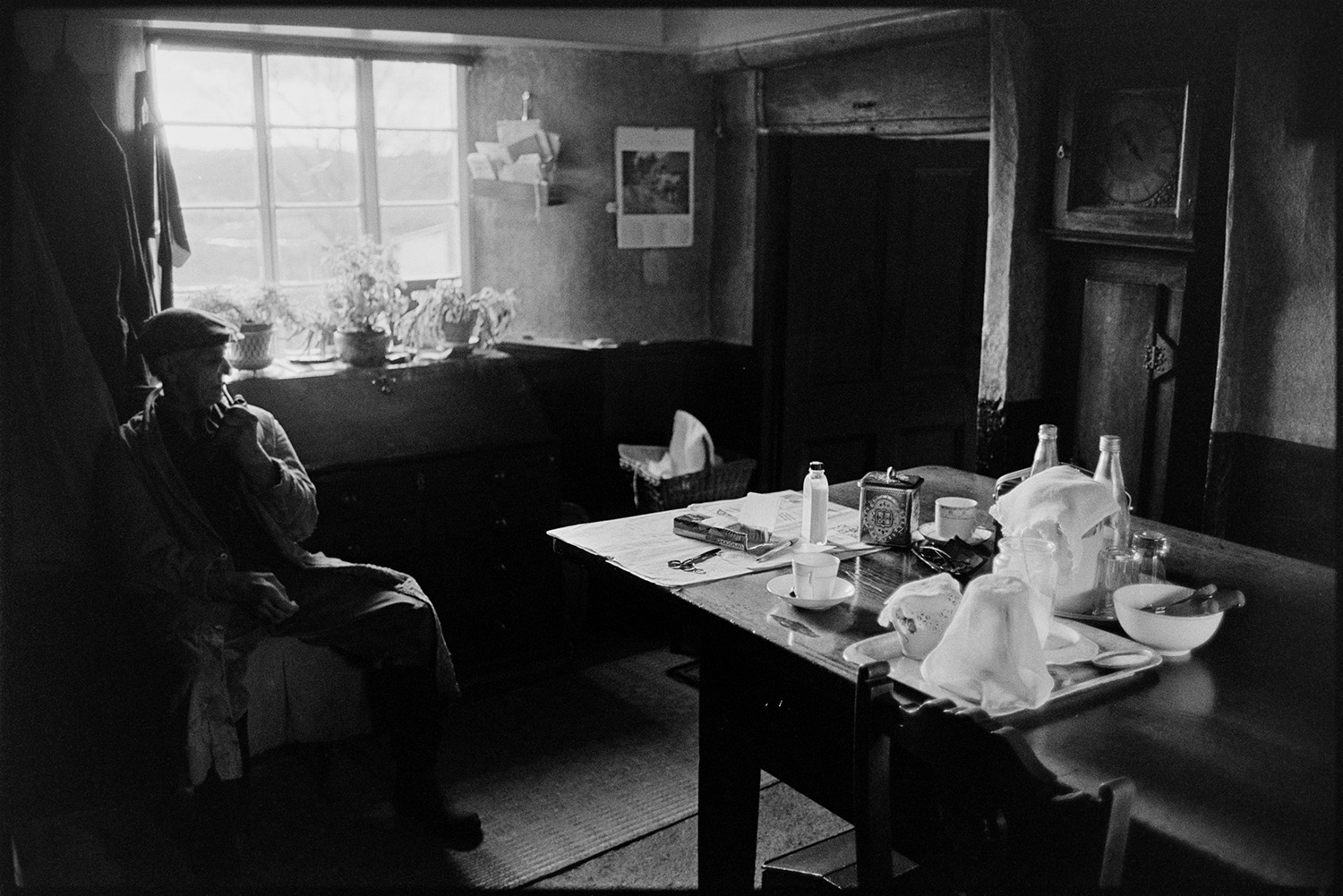 Farmhouse kitchen with tea on table. 
[Reg Chamings sat in the kitchen of Bridgetown farmhouse. The table has cups, a teapot, newspaper and pair of scissors on it. A grandfather clock can be seen in the background and the window sill has various pot plants.]