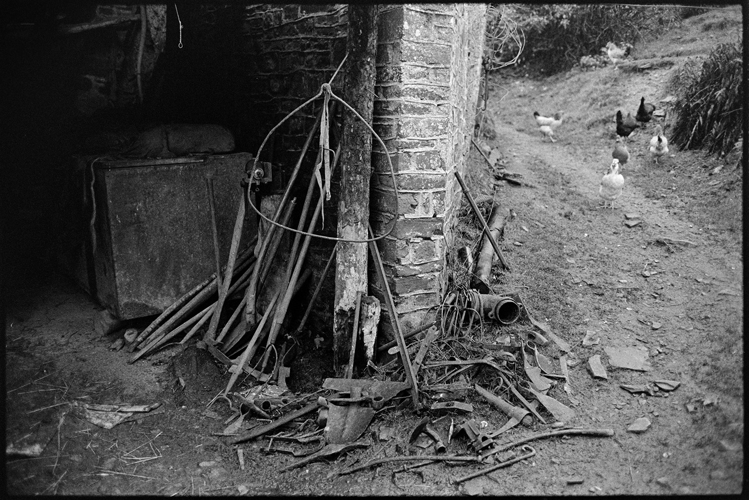 Tools and scrap in shed. <br />
[Items of scrap metal by a brick shed at Millhams in Dolton. A group of chickens are visible on a path running past the shed.]