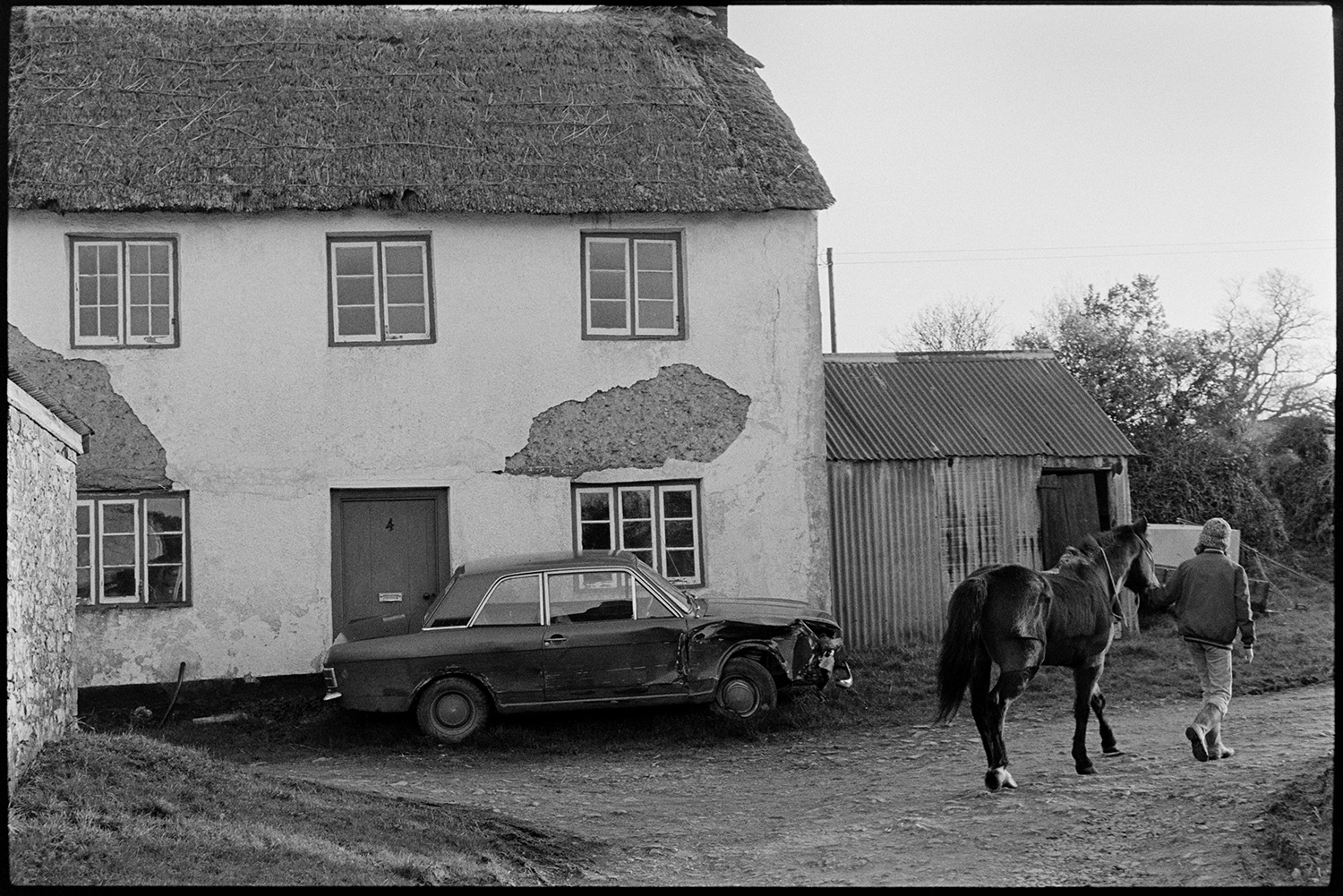 Cottages, thatch and cob, cottage sadly renovated and ruined!!<br />
[A person leading a horse past a thatch and cob house with a corrugated iron shed at High Bickington. A car with a dented bonnet is parked outside the house.]