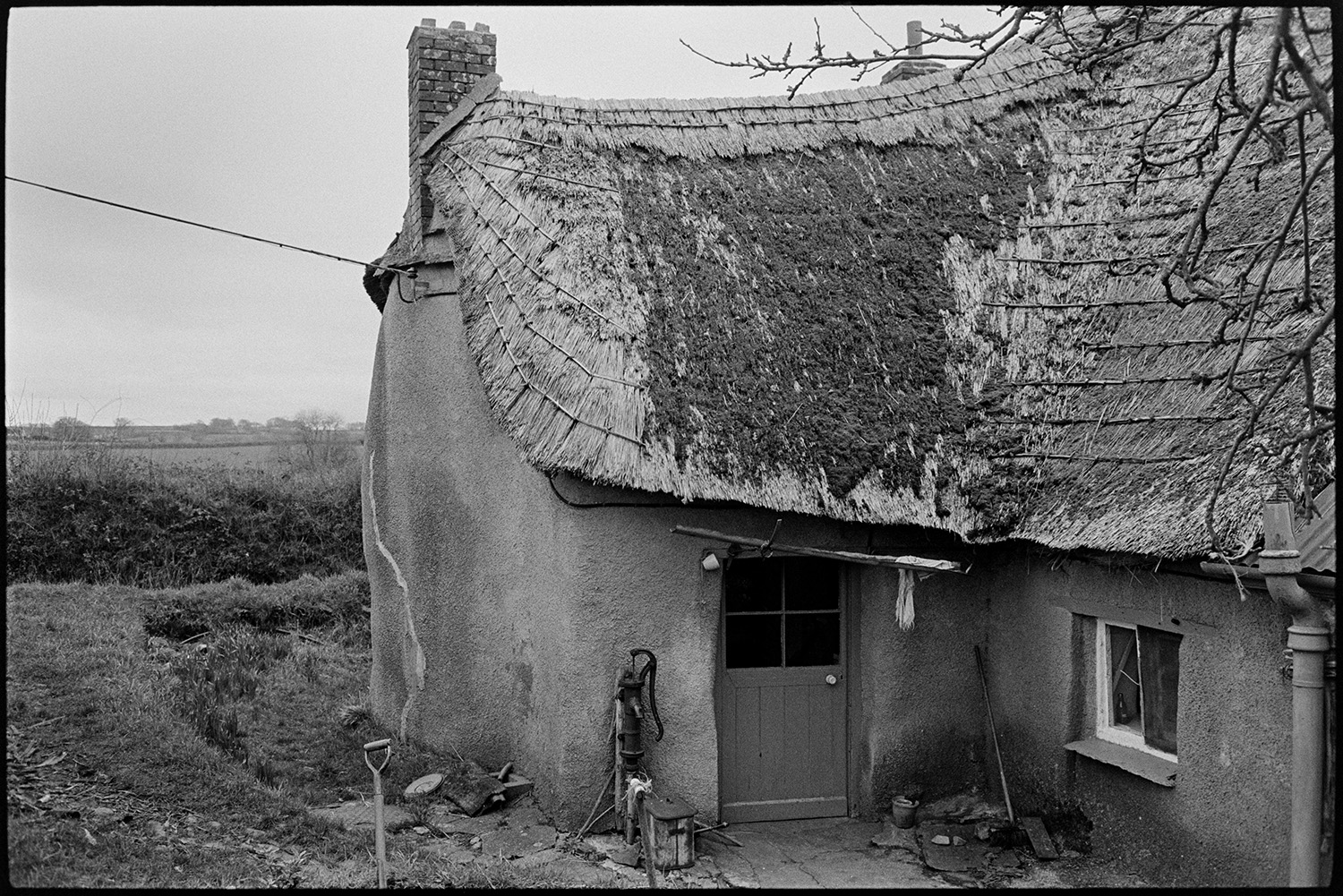 Cottage, cob and thatch, good interesting shape. 
[A cob and thatch cottage at Monkokehampton. The oddly shaped thatch roof is covered in moss and a pump is visible outside the front door.]