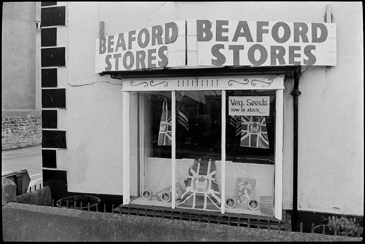 Front of village stores. 
[The shop front window of Beaford Stores in Beaford. Items for Queen Elizabeth II Silver Jubilee are displayed in the window, as well as a sign advertising vegetable seeds.]