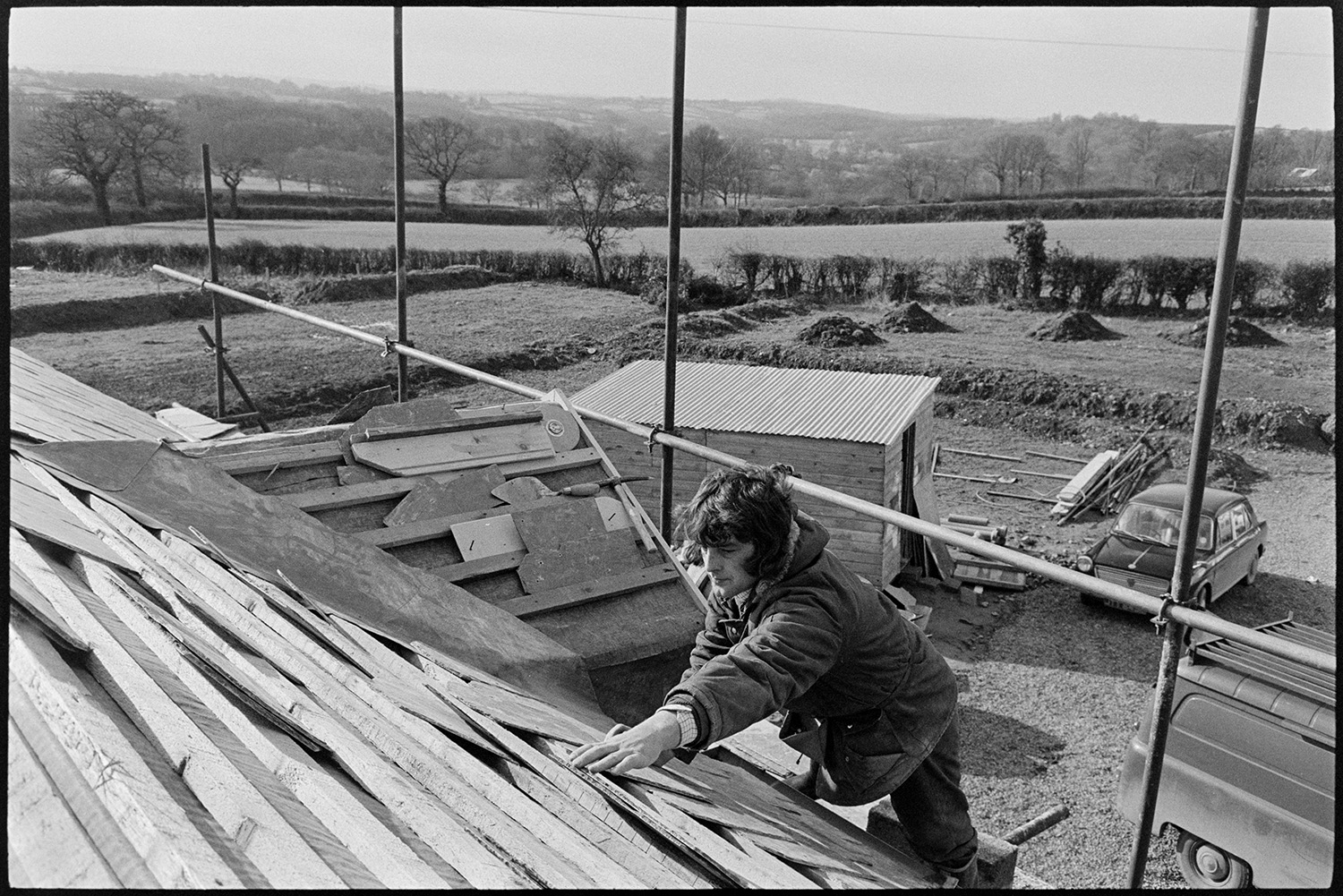 Man slating roof. <br />
[A man nailing slates to a roof at Langlands, Iddesleigh. He is stood on scaffolding. A car, van, wooden shed, fields, hedges and trees can be seen in the background.]