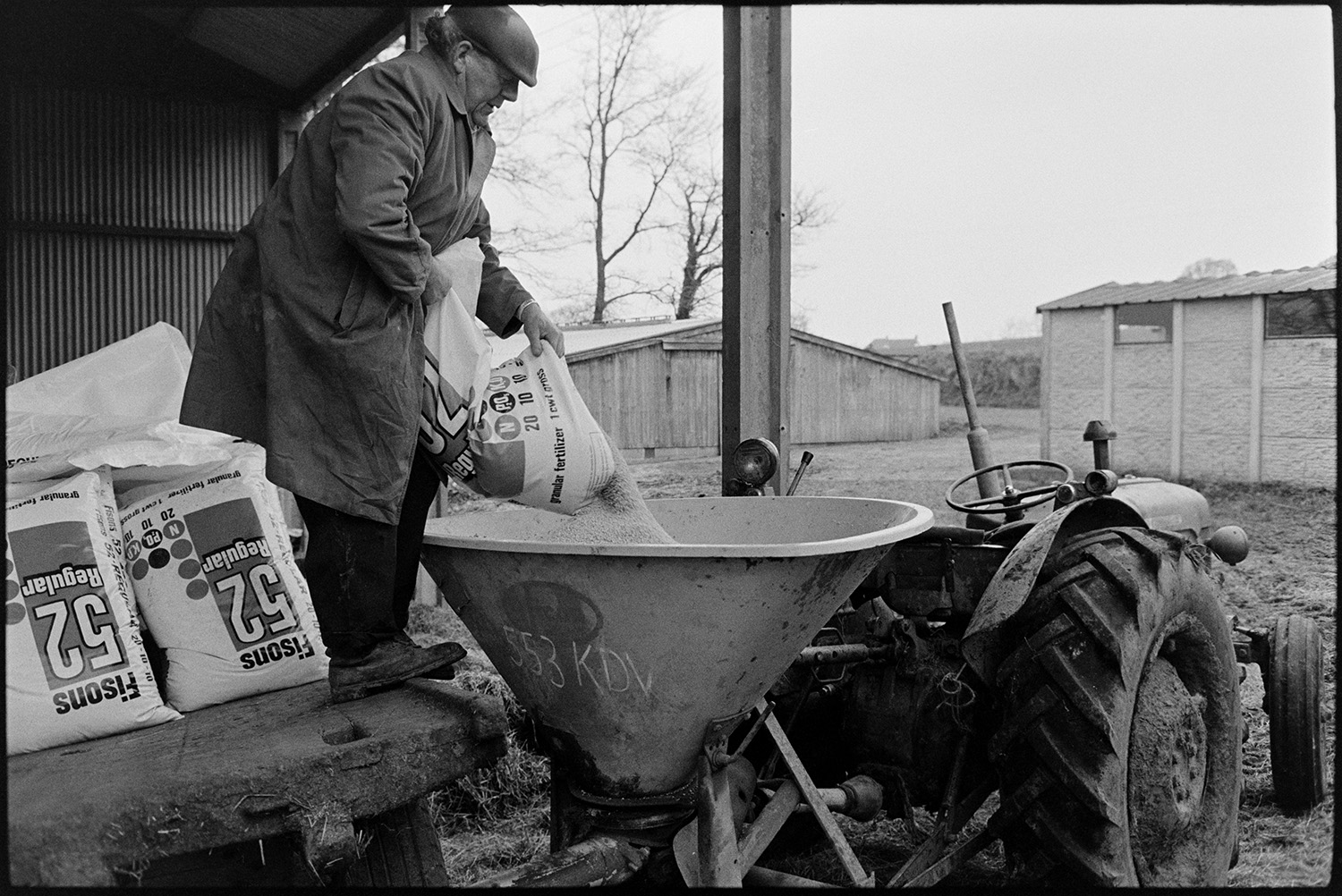 Man loading up and spreading fertilizer. 
[John Ward loading fertilizer into a funnel attached to a tractor in the farmyard, at Parsonage, Iddesleigh.]