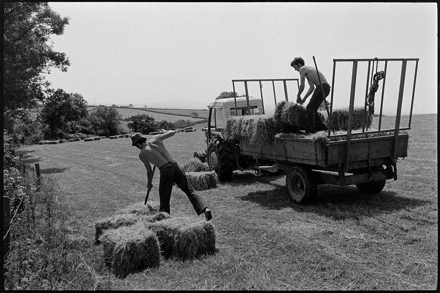 David Ward and Graham Ward loading hay bales onto a trailer in a field at Parsonage, Iddesleigh. One of the men is picking up a bale with a pitchfork. More bales can be seen in the field in the background.