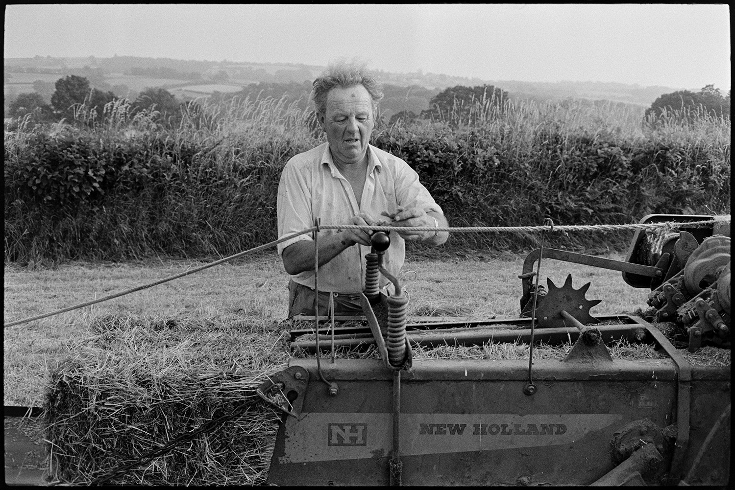 Farmers baling hay. 
[John Ward checking his baler in a field at Parsonage, Iddesleigh. A hay bale can be seen in the baler.]
