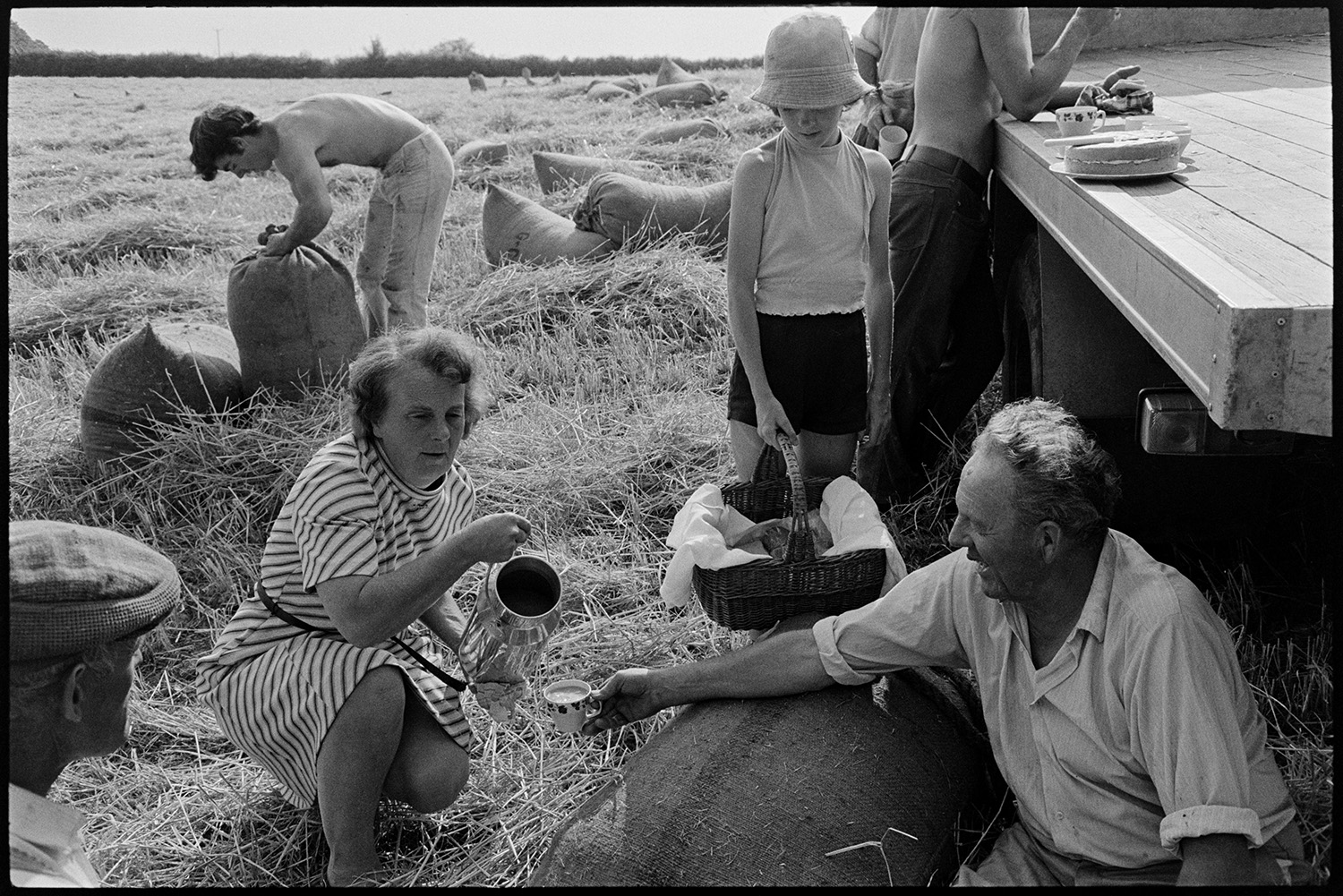 Harvesters tea break in field. 
[Hettie Ward bringing tea and cake to the Ward family who are harvesting in a field at Parsonage, Iddesleigh. She is pouring tea from an urn into a china cup held by John Ward. A girl is also stood holding a basket of food by them and a cake can be seen on the trailer behind them.]
