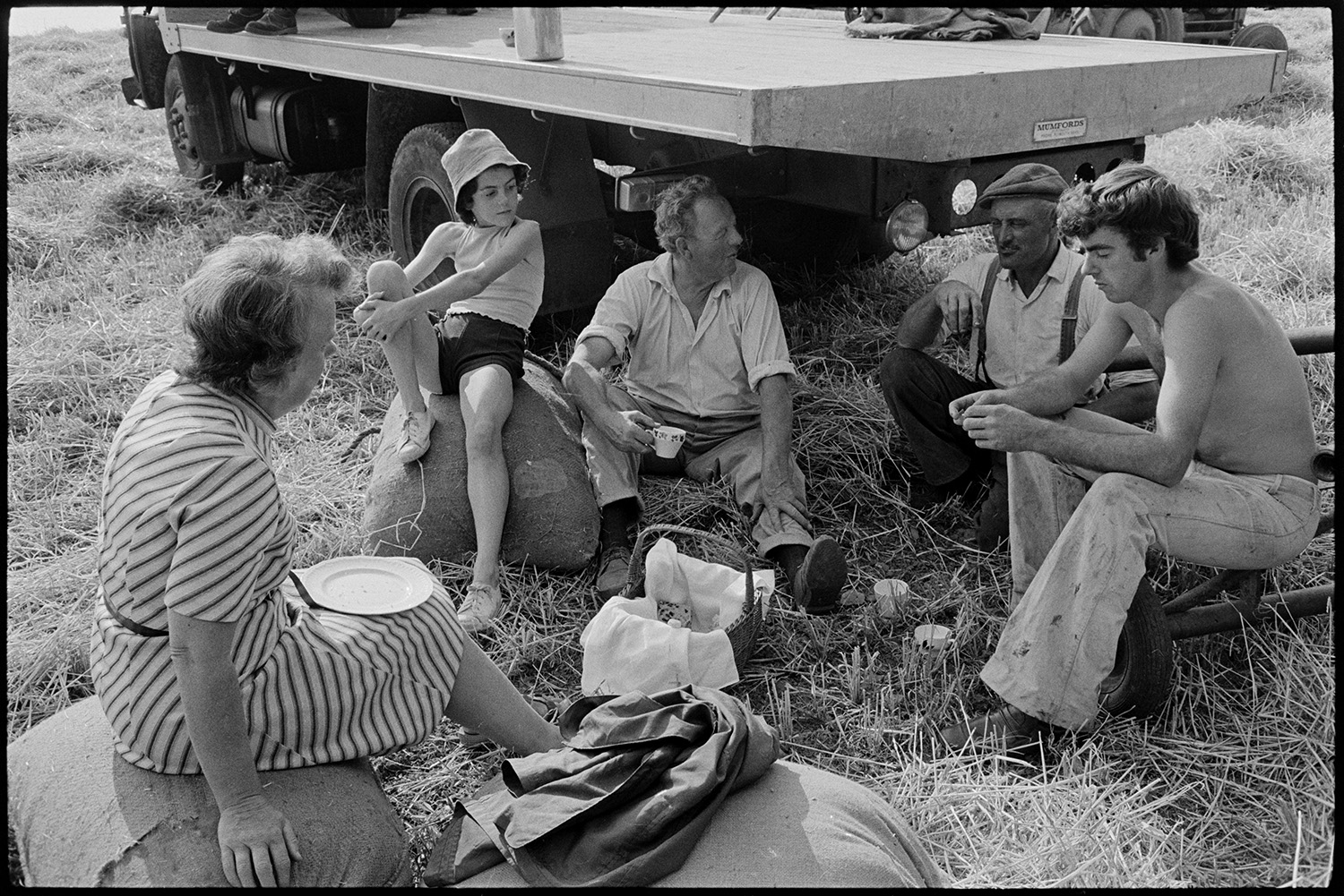 Harvesters tea break in field. 
[The Ward family having a tea break whilst harvesting in a field at Parsonage, Iddesleigh. Hettie Ward and a girl at sat on hessian sacks. John ward is sat on the ground with two other men.]