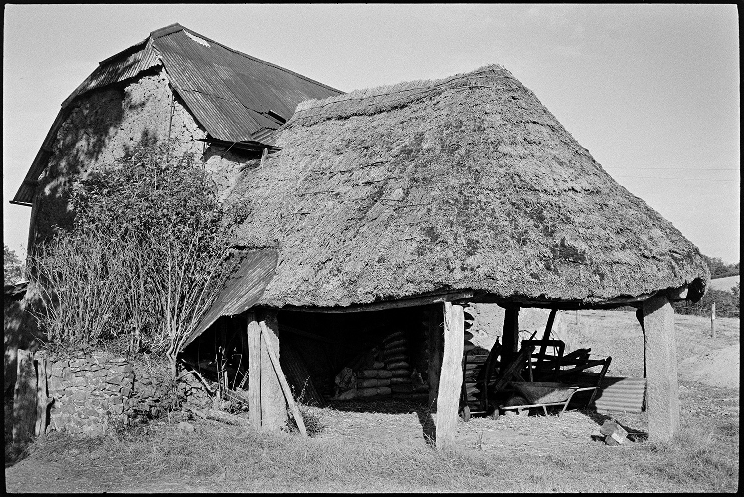 Barn and thatched roundhouse with pillars. 
[A cob barn with a corrugated iron roof and thatched roundhouse with wooden and stone pillars at Chapple, Winkleigh. A wheelbarrow and sacks can be seen under the roundhouse.]
