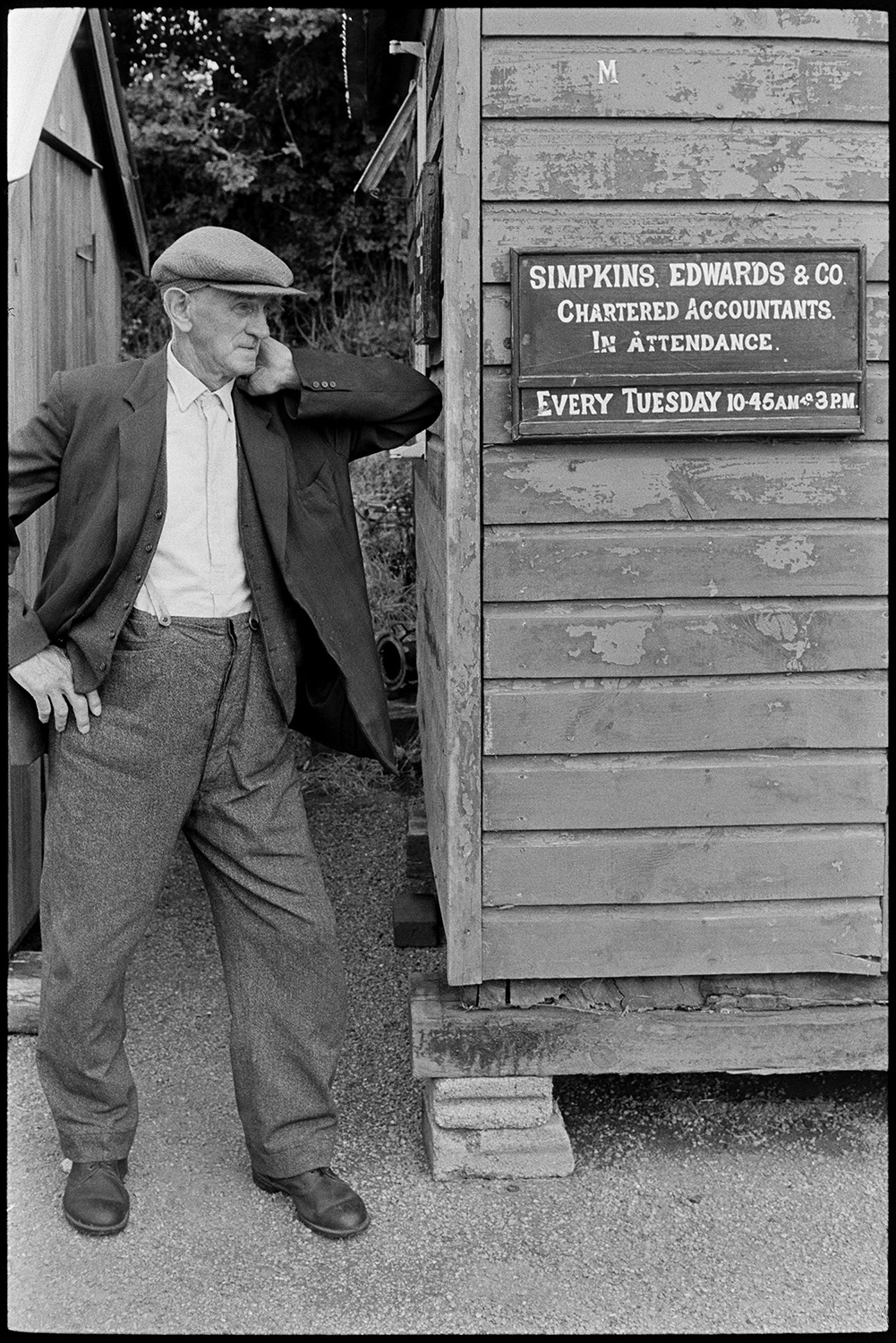 People around on market day, clothes.
[A man wearing a flat cap leaning against a wooden shed at Hatherleigh Market. A sign on the shed reads 'Simpkins, Edward & Co. Chartered Accountants. In Attendance Every Tuesday 10-45AM to 3PM.'.]