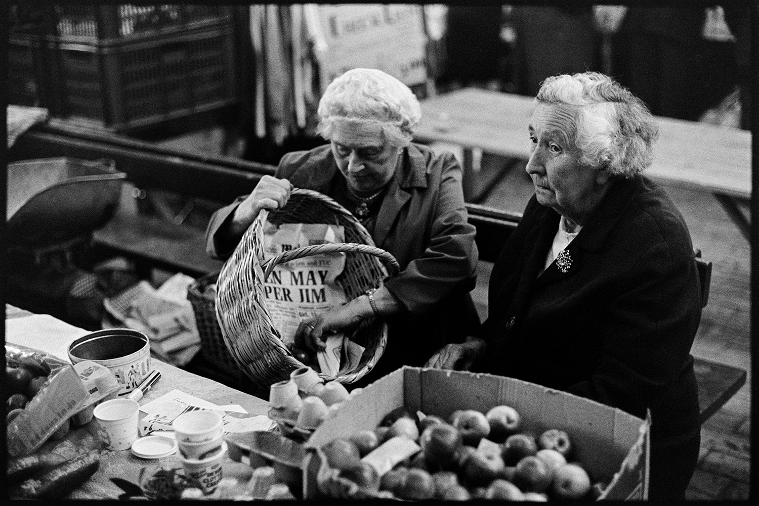 Stalls at Pannier Market, food, clothes. 
[Two women sat behind a stall at Bideford Pannier Market. One lady is emptying produce from a wicker basket. Eggs and apples are displayed on the stall for sale.]