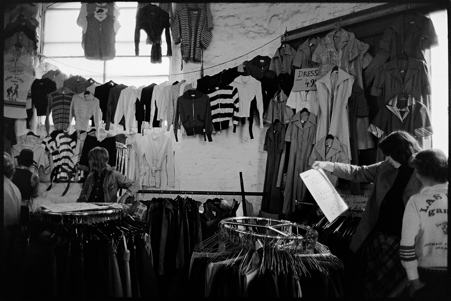 Stalls at Pannier Market, food, clothes. 
[People looking at items on a clothes stall in Bideford Pannier Market. The clothes are displayed on rails and strung across the wall of the pannier market.]