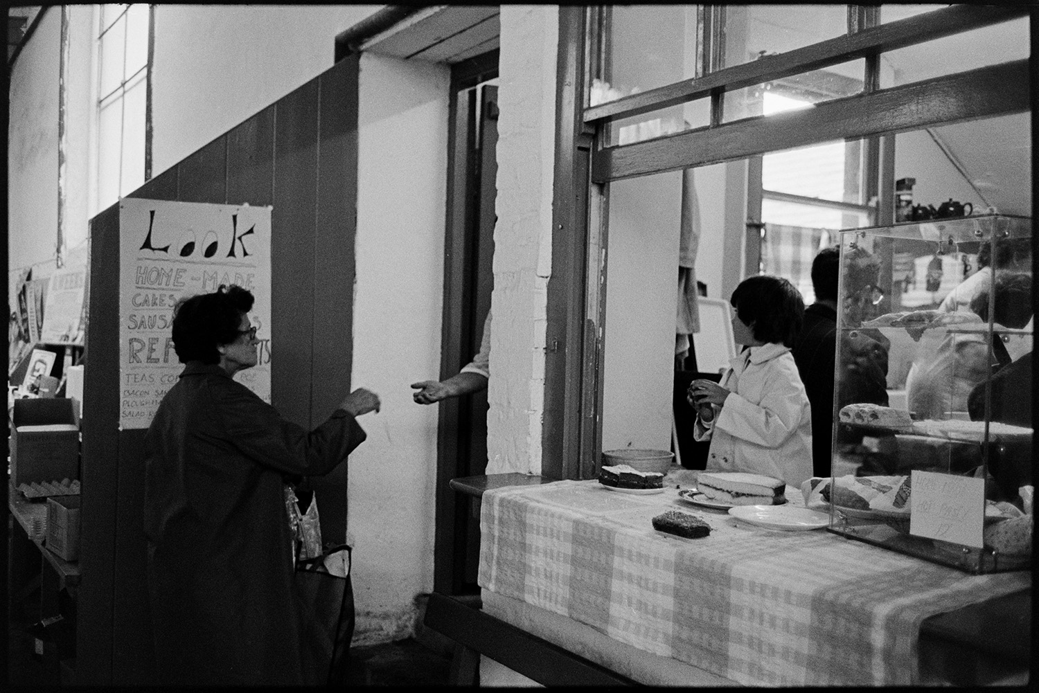 Stalls at Pannier Market, food, clothes. 
[A woman paying for cakes at a shop doorway in Bideford Pannier Market. A child is stood behind the shop hatch and cakes are on display.]