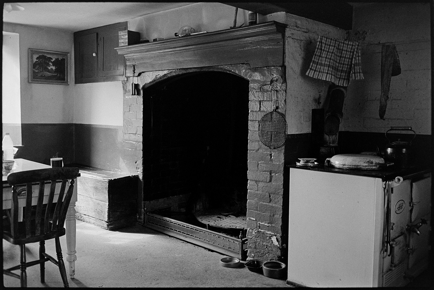 Fireplace and Rayburn stove in farmhouse. 
[The kitchen in a farmhouse at West Chapple, Winkleigh. An open fireplace and rayburn stove are visible, as well as a dining table and chair. A picture of a landscape scene is hung on the wall and a ship in a bottle is displayed on the mantelpiece.]