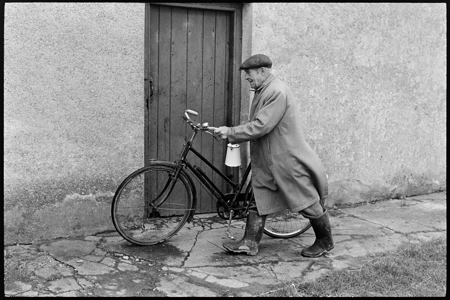 Man with bicycle. 
[A man wearing wellington boots and pushing a bicycle past a wooden door.]