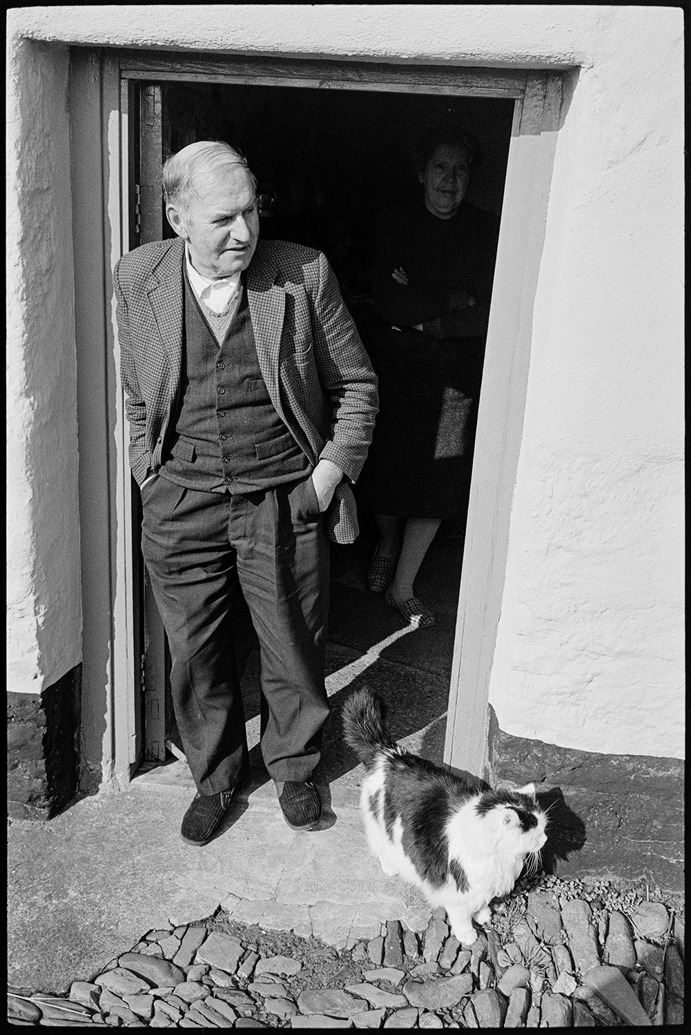 Woman and man at door with cat. 
[Mr Vanstone stood in his doorway with his cat at Monkokehampton. The pathway outside the house is cobbled. Mrs Vanstone can be seen inside the house.]