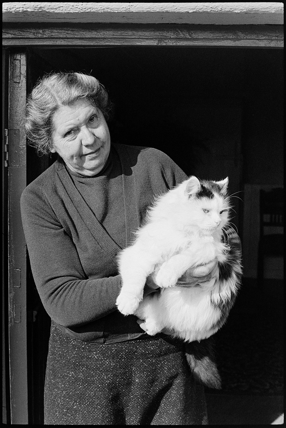 Woman and man at door with cat. 
[Mrs Vanstone stood in her doorway holding her cat, at Monkokehampton.]