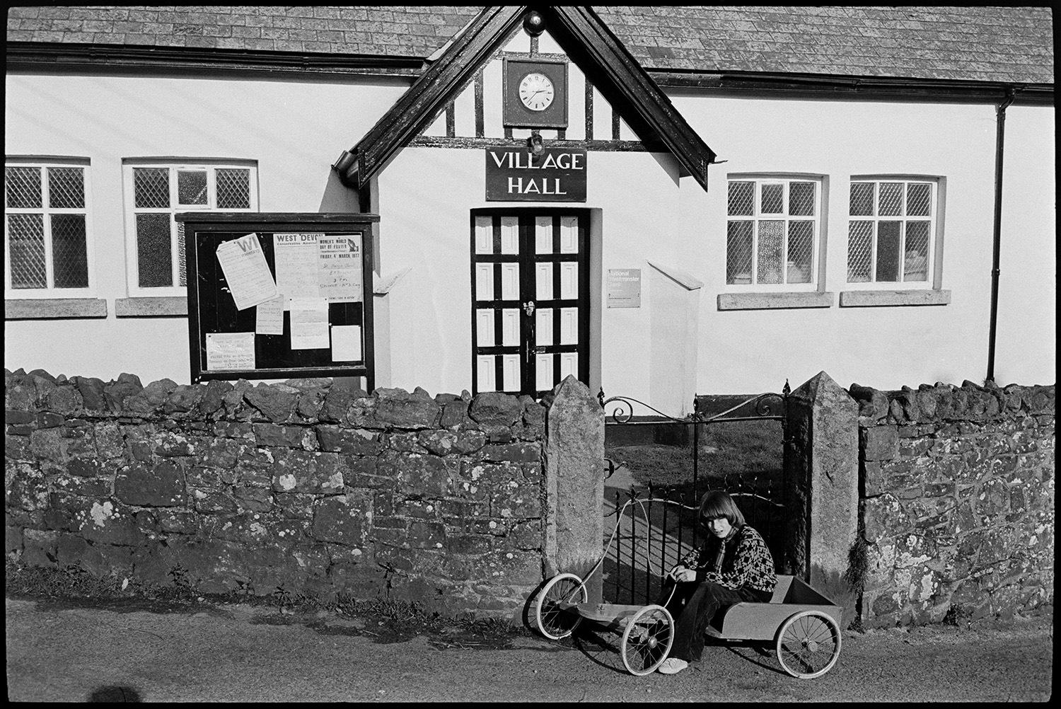 Village Hall. 
[Kings Nympton Village Hall. A child on a soap box is outside the gate to the hall. A clock and notice board at next to the hall entrance.]