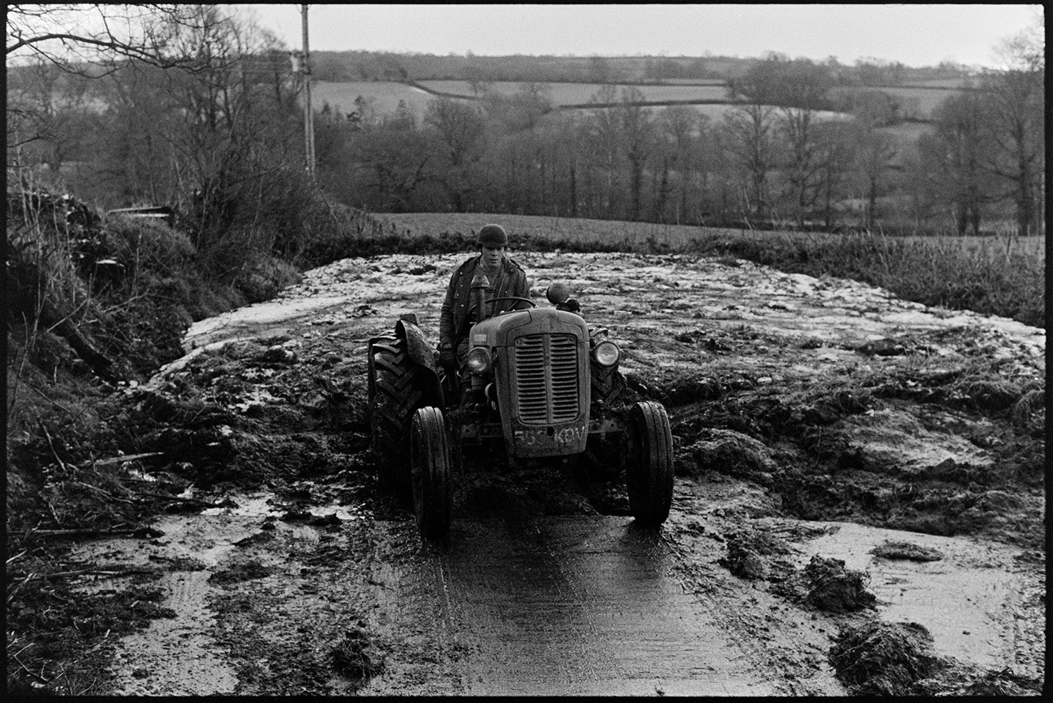 Tractor and scraper in mucky yard. 
[David Ward using a tractor and scraper to clear muck into a pile in a yard at Parsonage, Iddesleigh. Trees and fields can be seen in the background.]