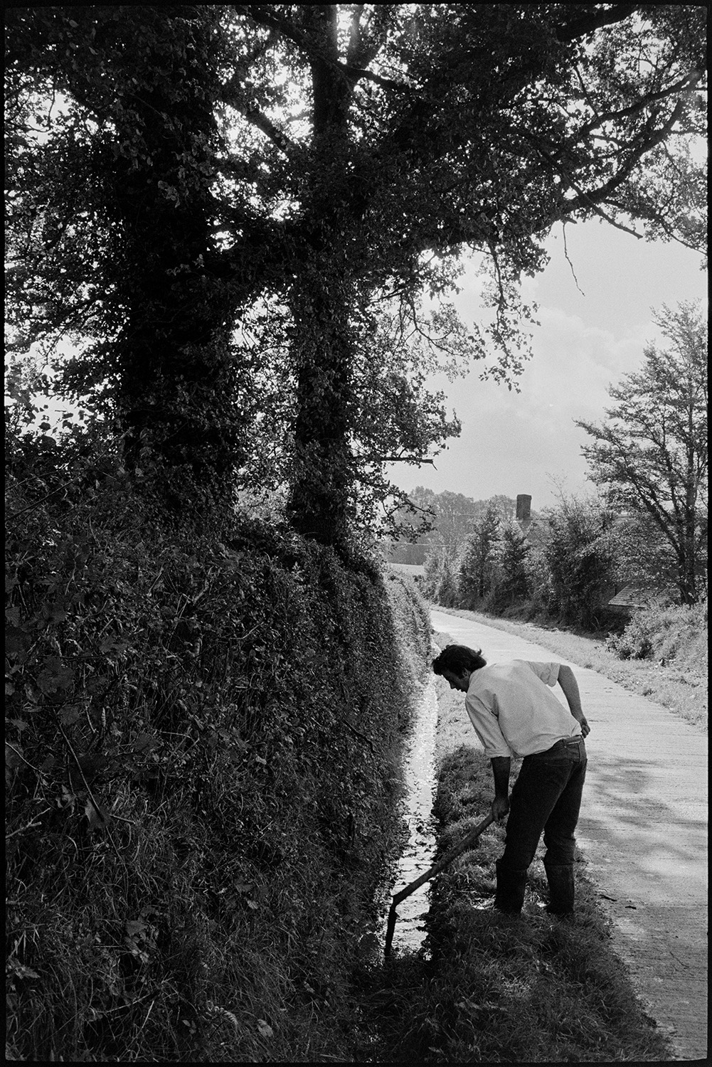 Man clearing ditch beside road. 
[Graham Ward clearing a ditch using a spade or shovel, by a roadside at Parsonage. Iddesleigh.]
