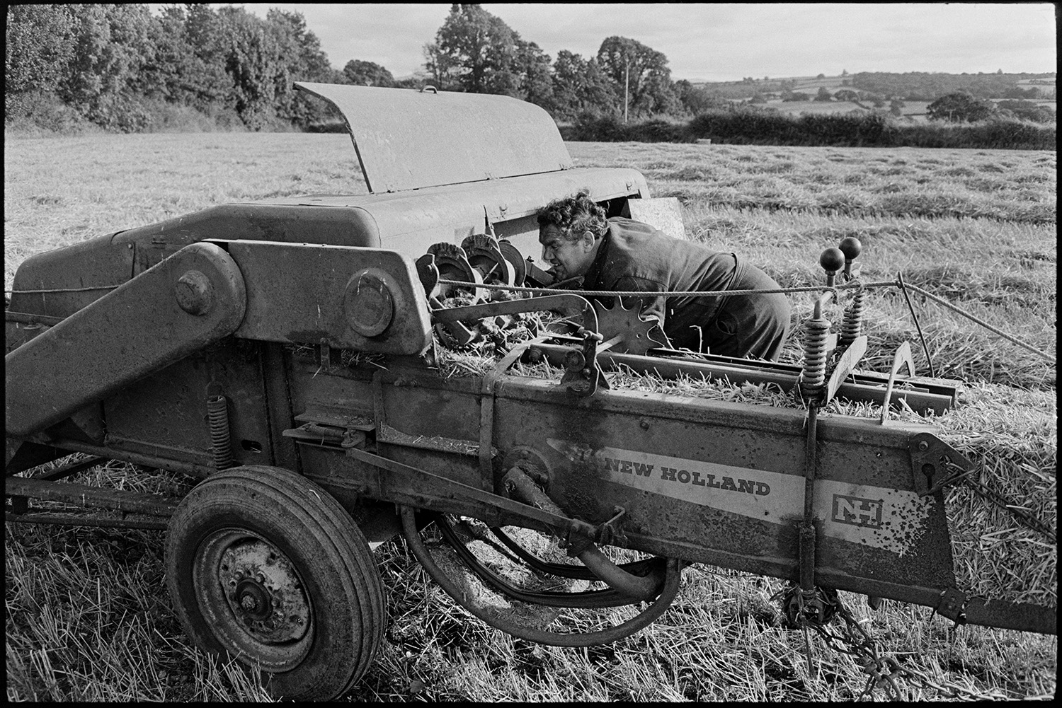 Mechanic checking baler. 
[Cyril Dunn checking the mechanics of a baler in a field at Parsonage, Iddesleigh. A bale of hay can be seen in the baler.]