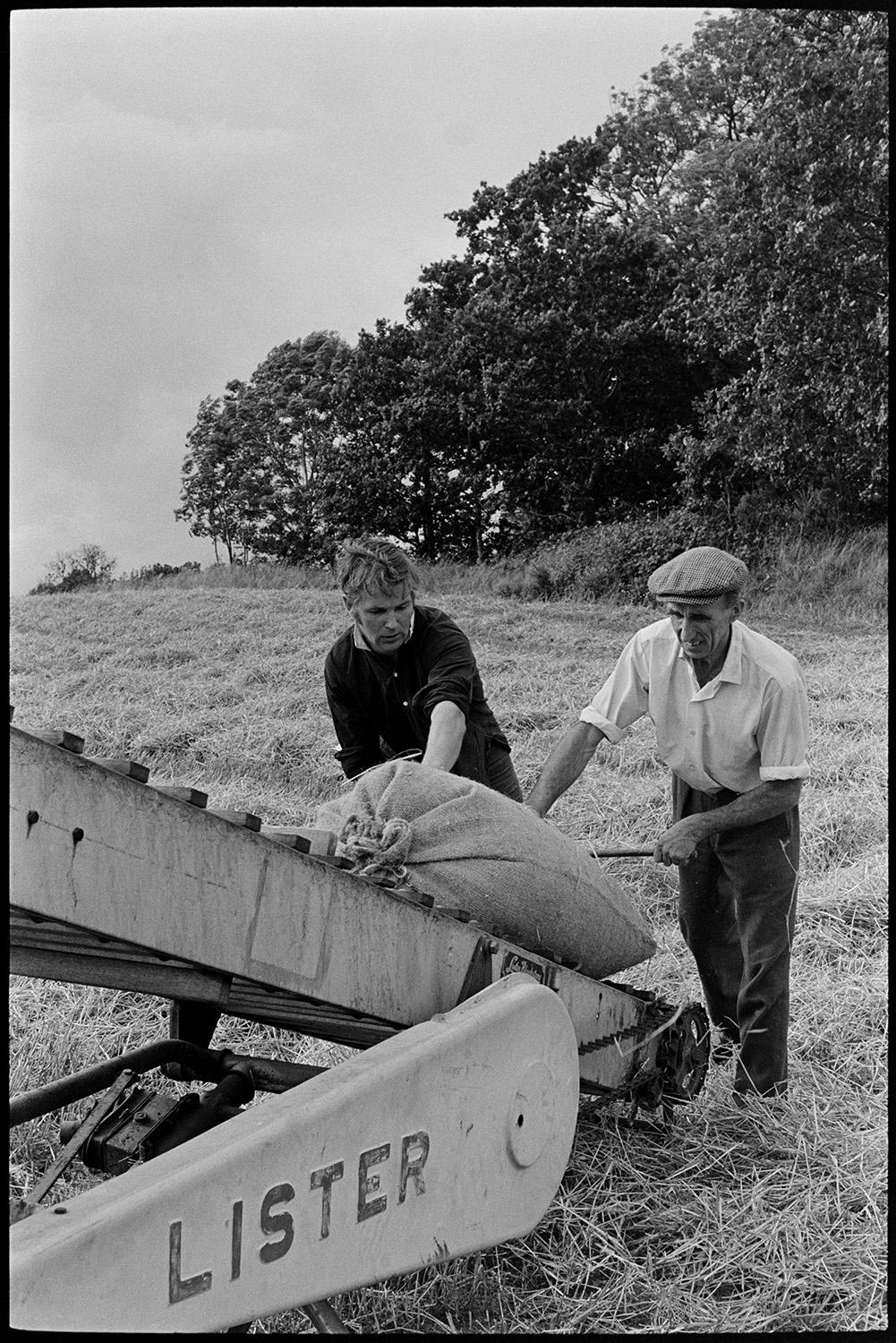Loading sacks of grain on to elevator. 
[Two men using an elevator to load sacks of grain onto a trailer in a field at Parsonage, Iddesleigh.]